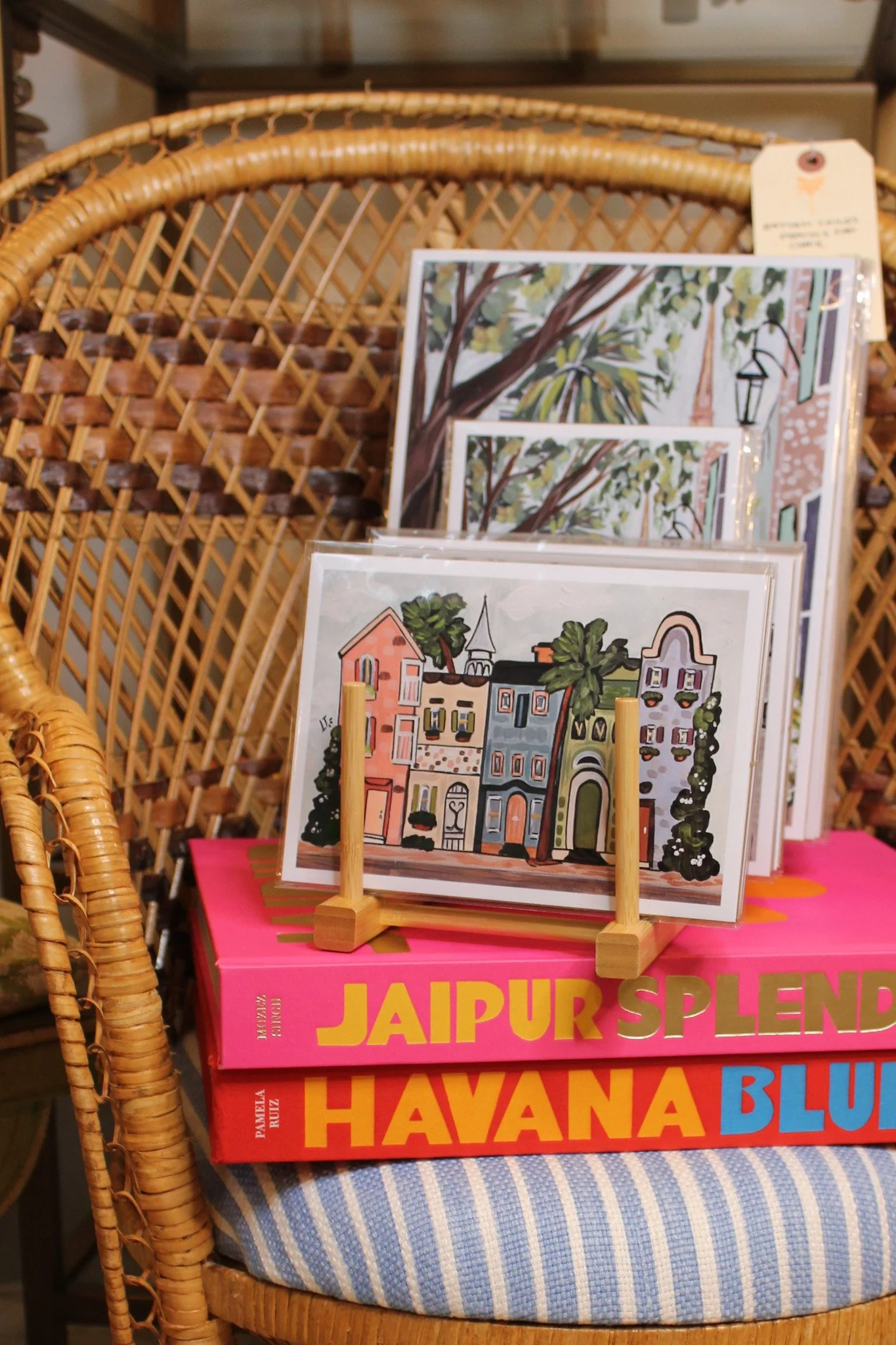 A display of colorful postcards with illustrations of buildings and trees, standing on a wooden rack in front of a rattan chair, with large brightly colored books titled 'Jaipur Splend' and 'Havana Blue' underneath.