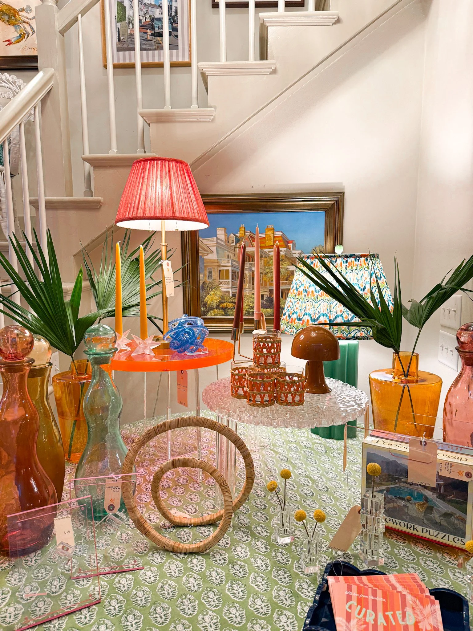 Display of colorful home decor items, including vases, candlesticks, lamps, and framed artwork on a table covered with a patterned cloth, with a staircase and framed photo in the background.