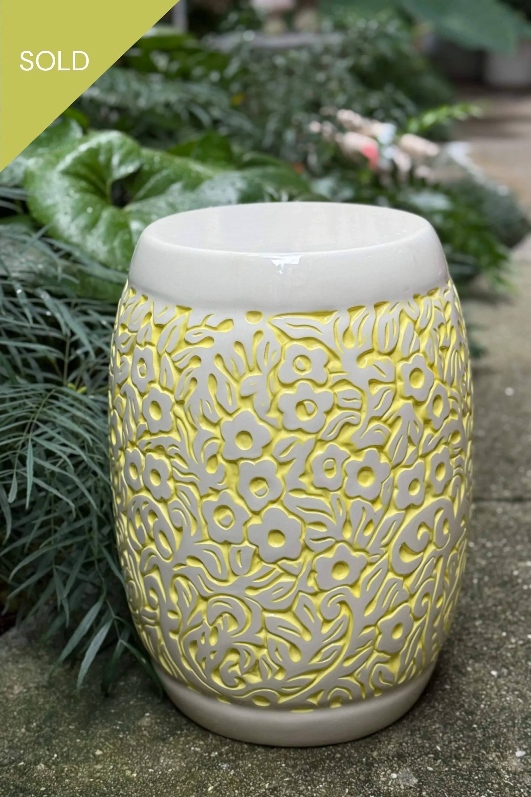 Decorative white ceramic stool with yellow floral pattern, placed outdoors on a concrete surface with green foliage in the background.