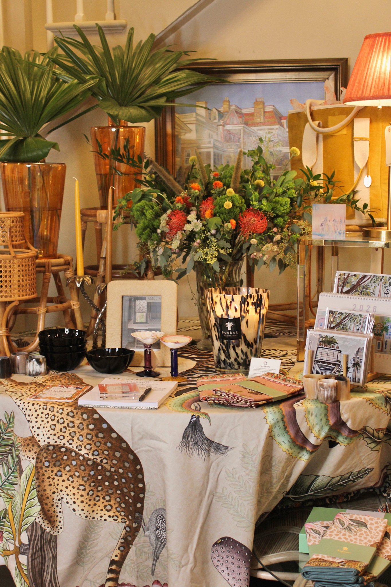A display table decorated with a jungle-themed tablecloth featuring animal prints, topped with various decorative items including vases, candles, framed pictures, notebooks, and a large bouquet of flowers and greenery. The background includes tall va