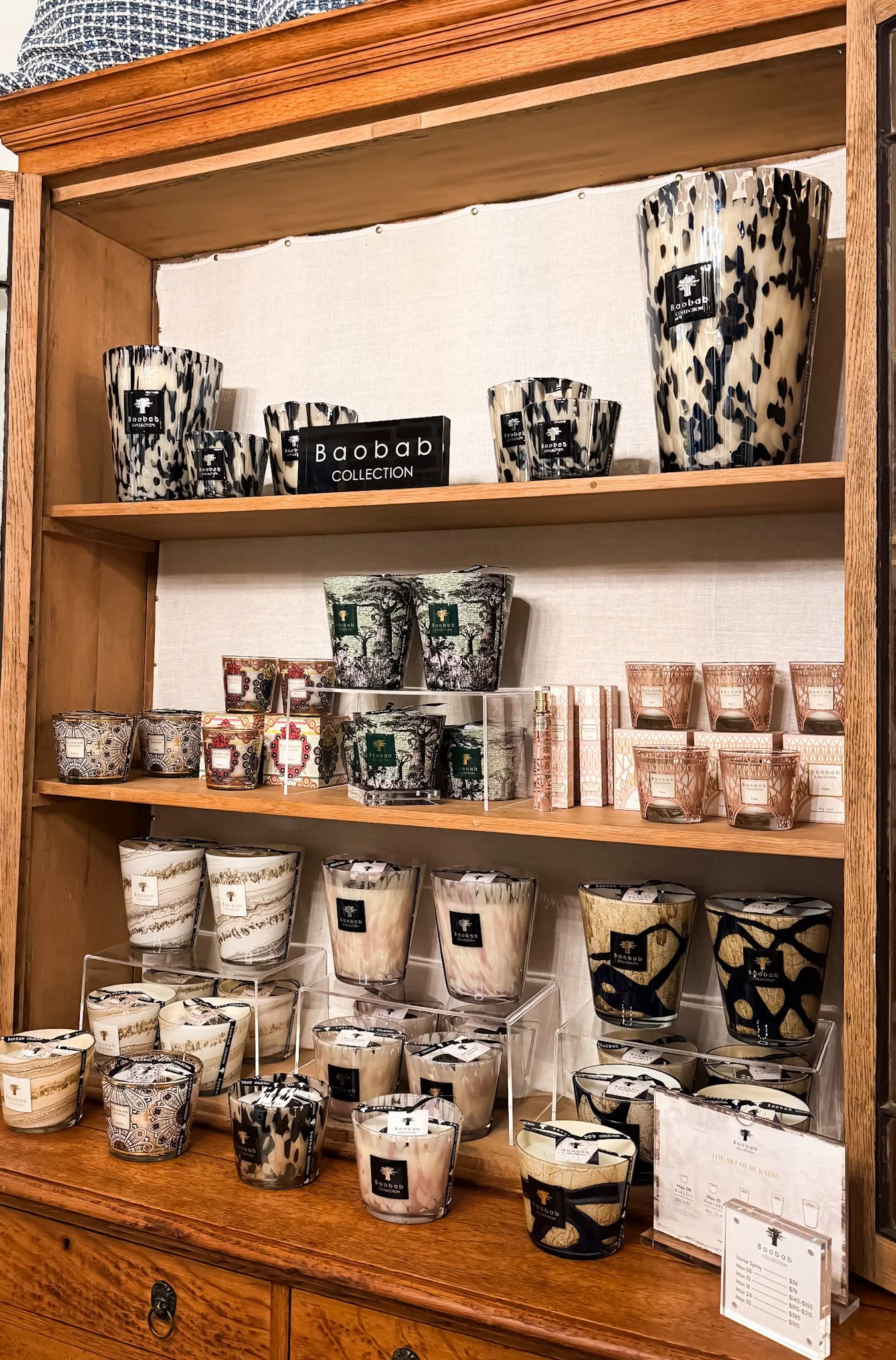 Display of decorative candles arranged on wooden shelves, featuring various patterns and sizes, with a sign reading 'Baobab COLLECTION'.