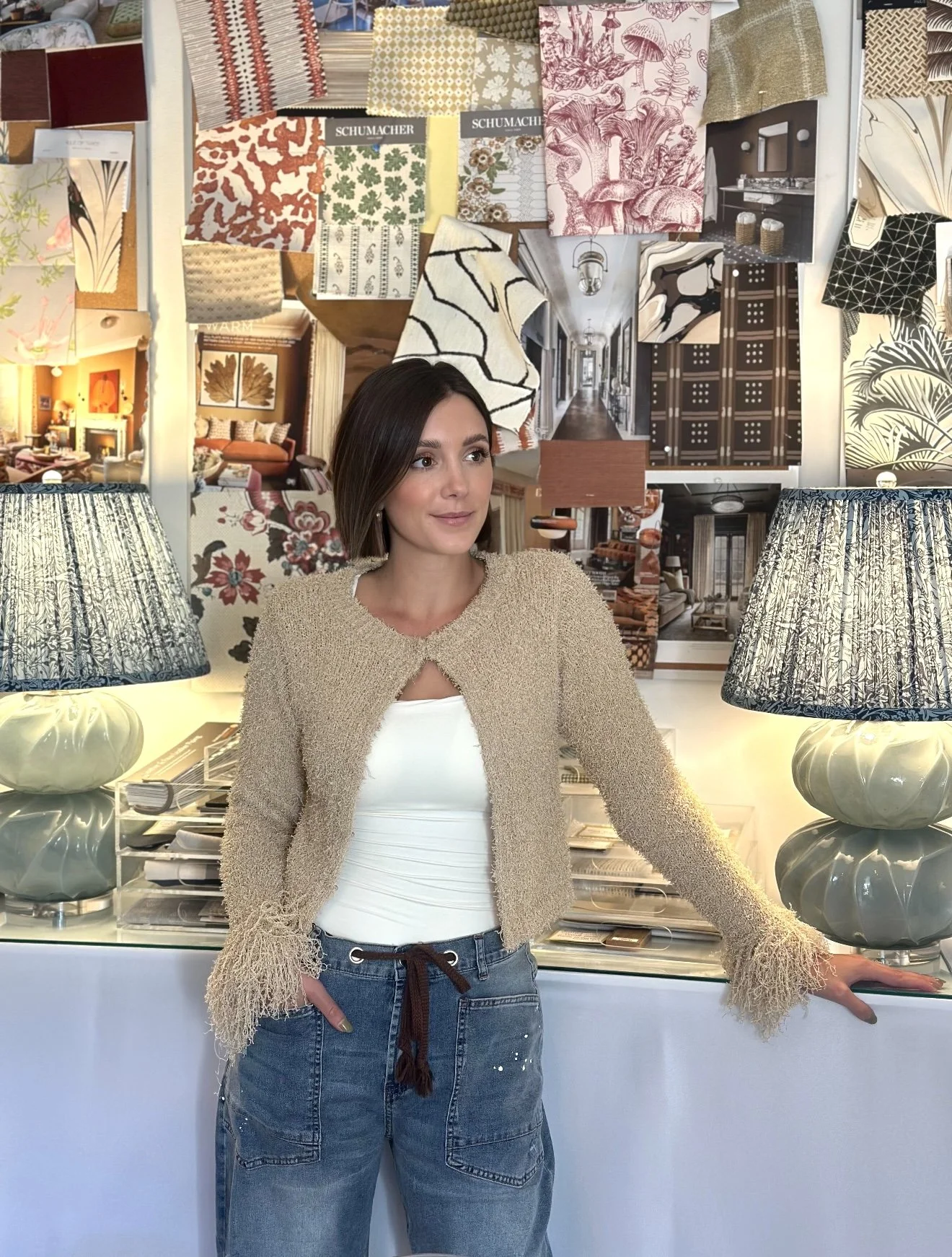A young woman with shoulder-length brown hair standing in a decor store, surrounded by fabric samples and decorative objects, with two lamps on a table behind her.