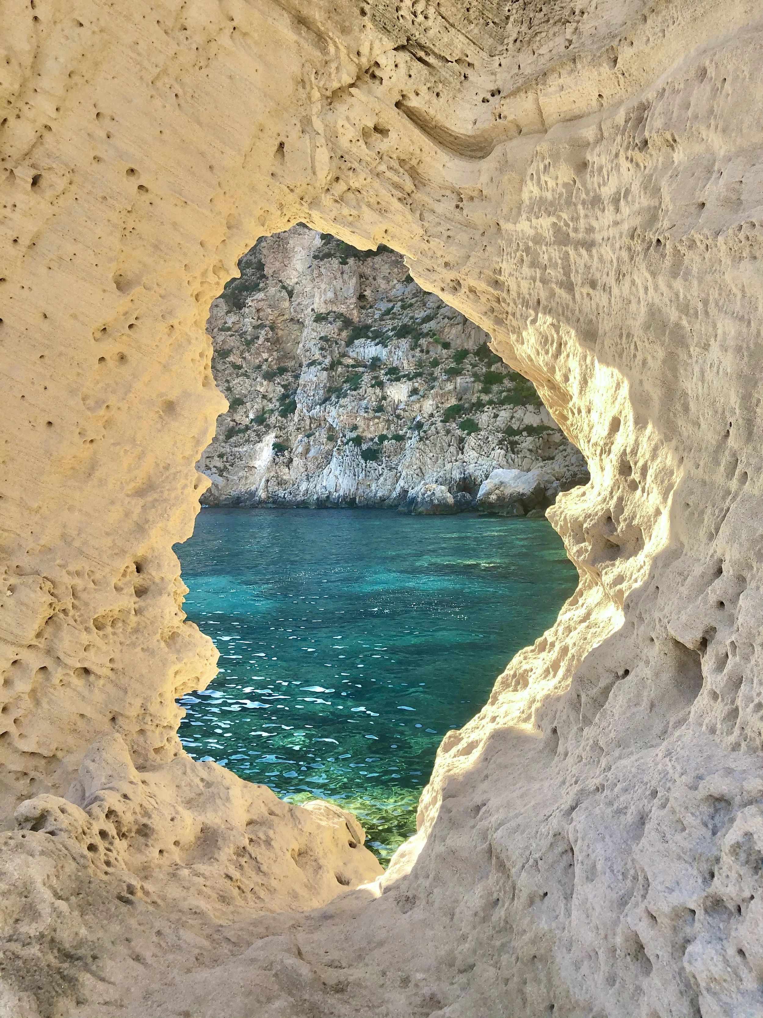 A view of the ocean through a natural rock formation with a smooth, rounded opening, overlooking a rocky cliffside with sparse green vegetation.