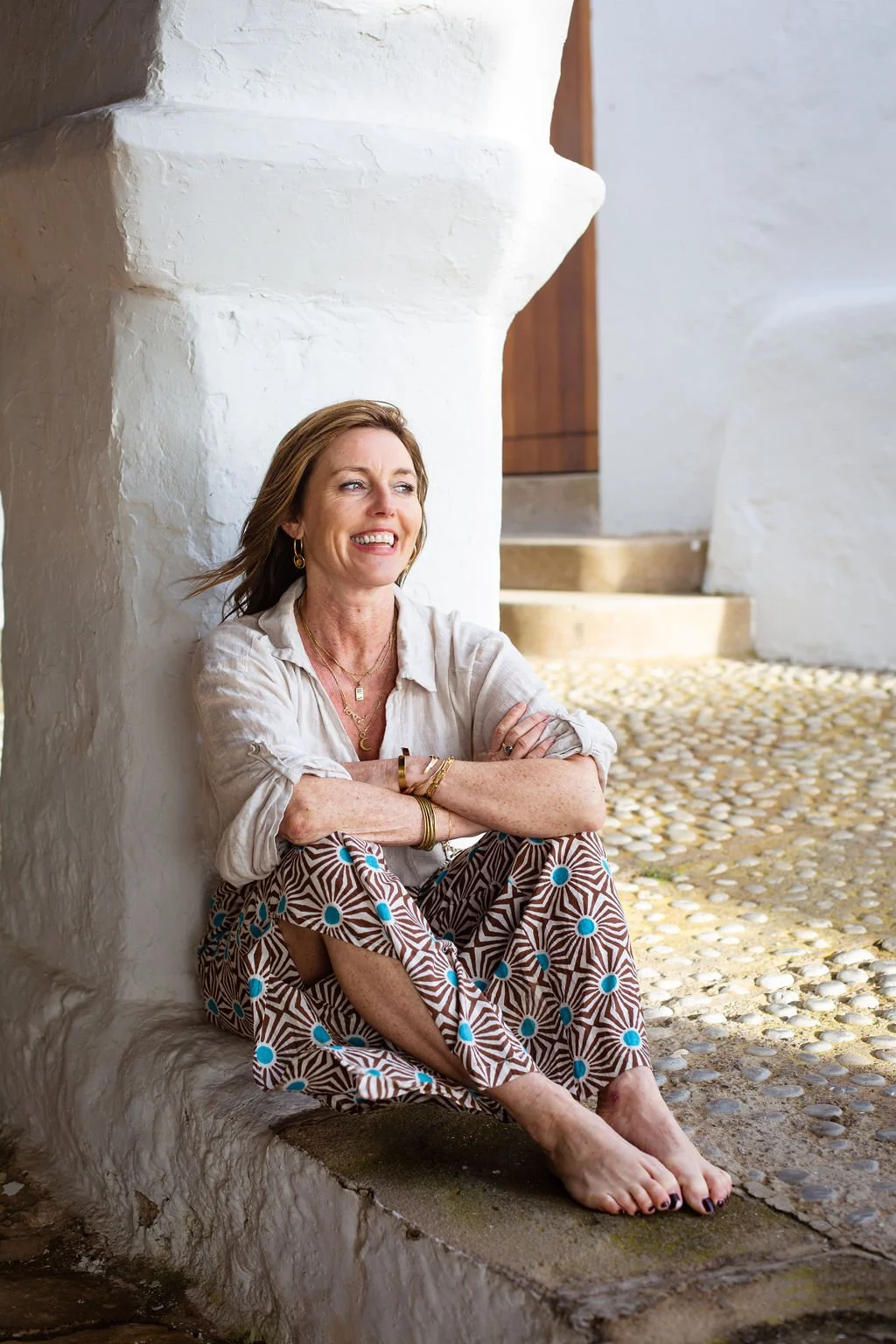 A woman sits on a stone ledge, leaning against a white textured wall with her arms crossed, smiling, with a staircase and a wooden door in the background.