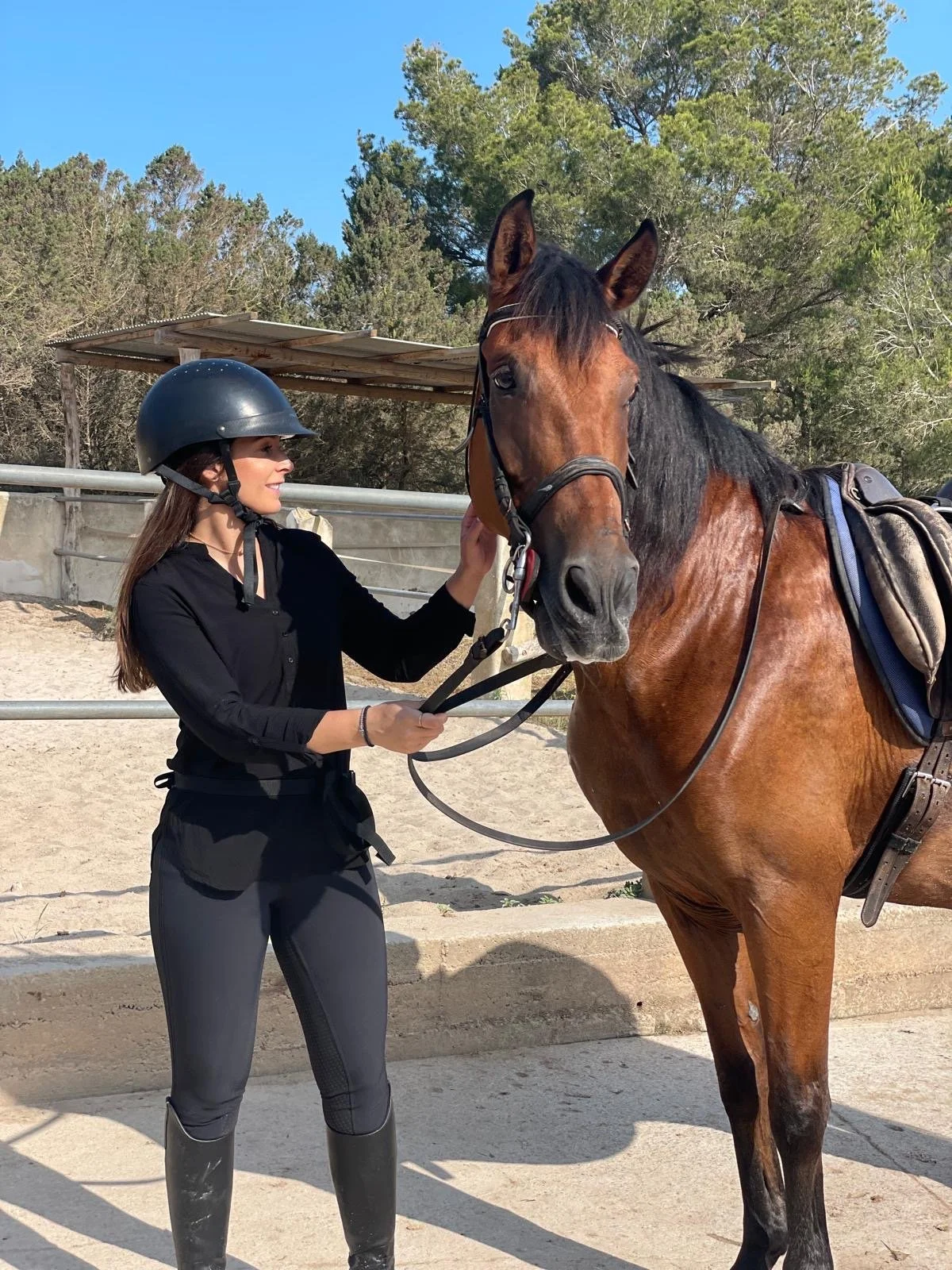 A woman wearing a black riding helmet, black long-sleeved shirt, and riding pants stands next to a brown horse with a black mane, holding its reins and touching its face. The scene is outdoors at a horse riding arena with a wooden shelter and trees i