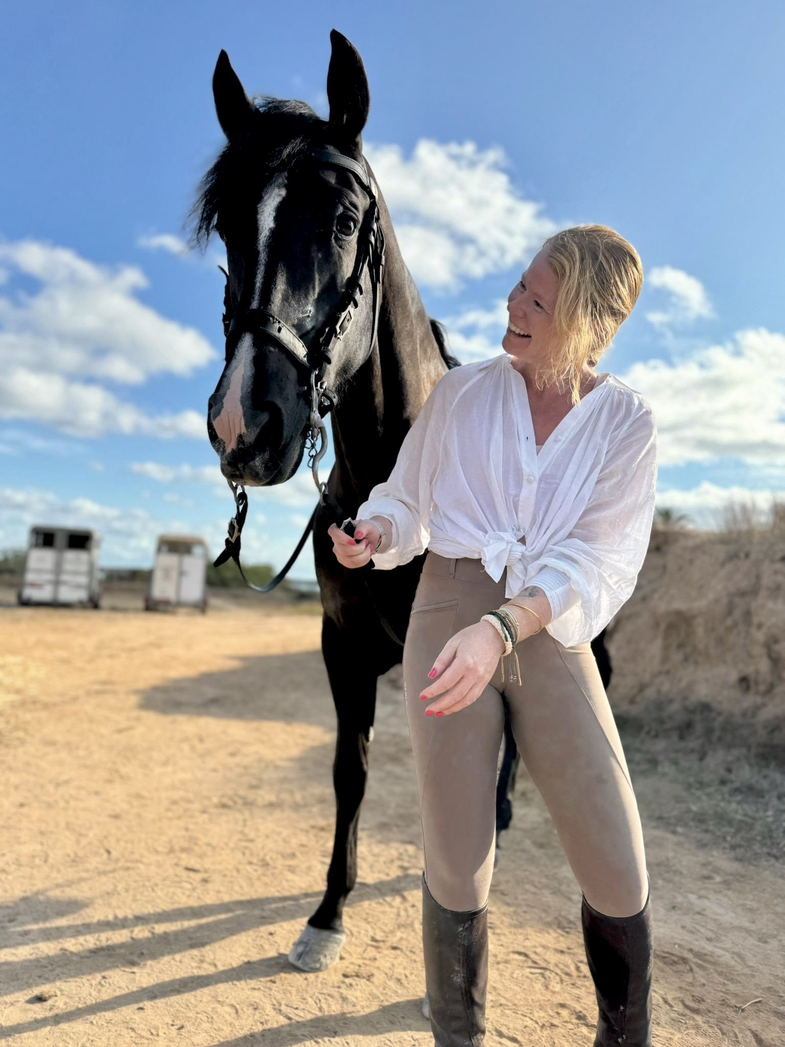 A woman with blonde hair, wearing a white blouse and beige riding pants, stands smiling next to a black horse on a sandy outdoor area under a blue sky with scattered clouds.