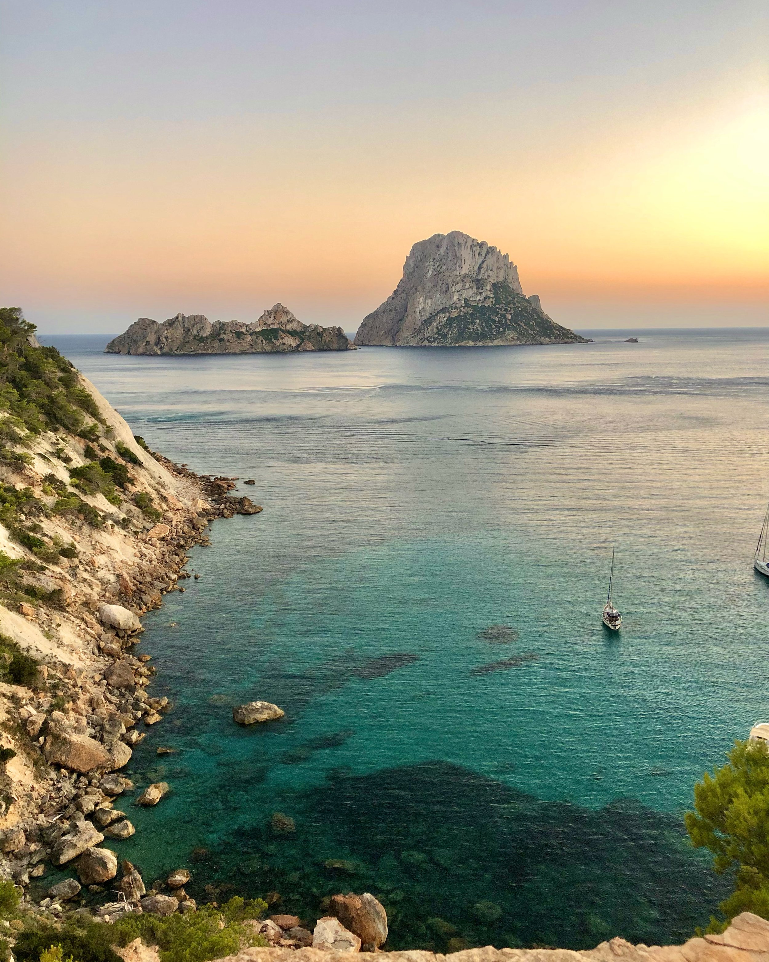 Scenic view of a coastal landscape at sunset with rocky cliffs, turquoise water with rocks, and sailboats, and large rock formations in the distance.