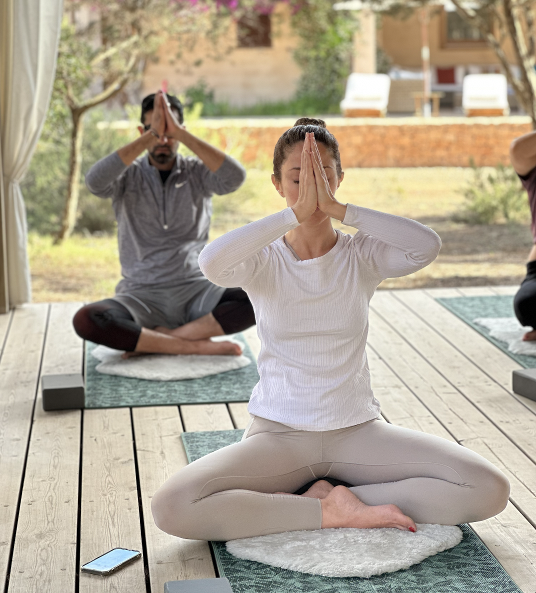 Three people practicing yoga outdoors on a wooden deck, seated in a meditative pose with hands together in prayer position.