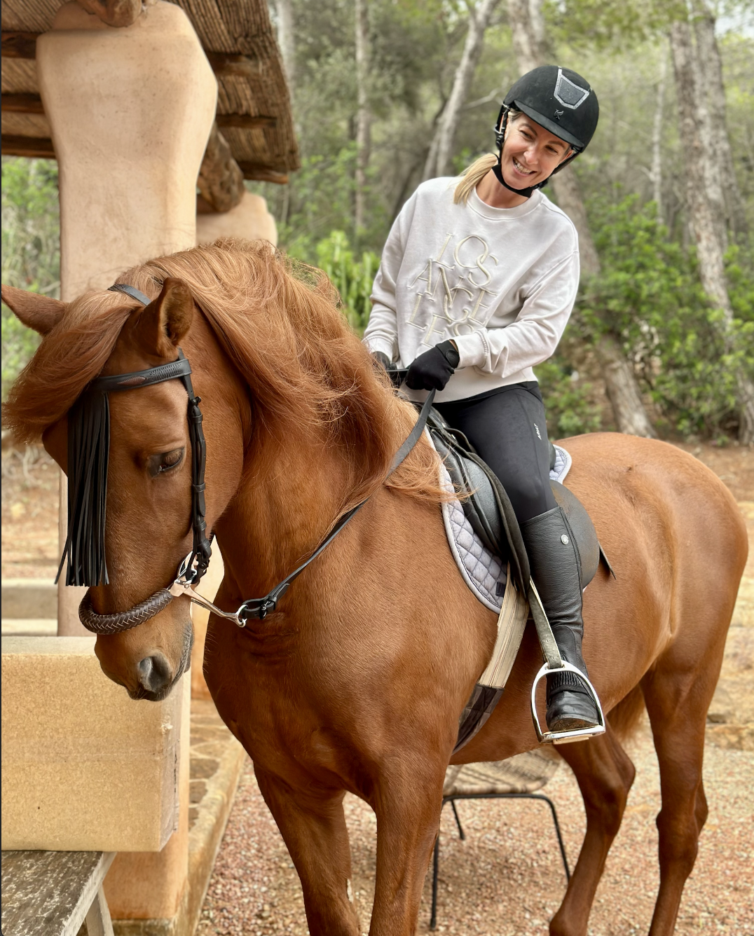 A woman riding a chestnut horse outdoors. She is wearing a helmet, white sweatshirt, black riding pants, and gloves, smiling at the horse.