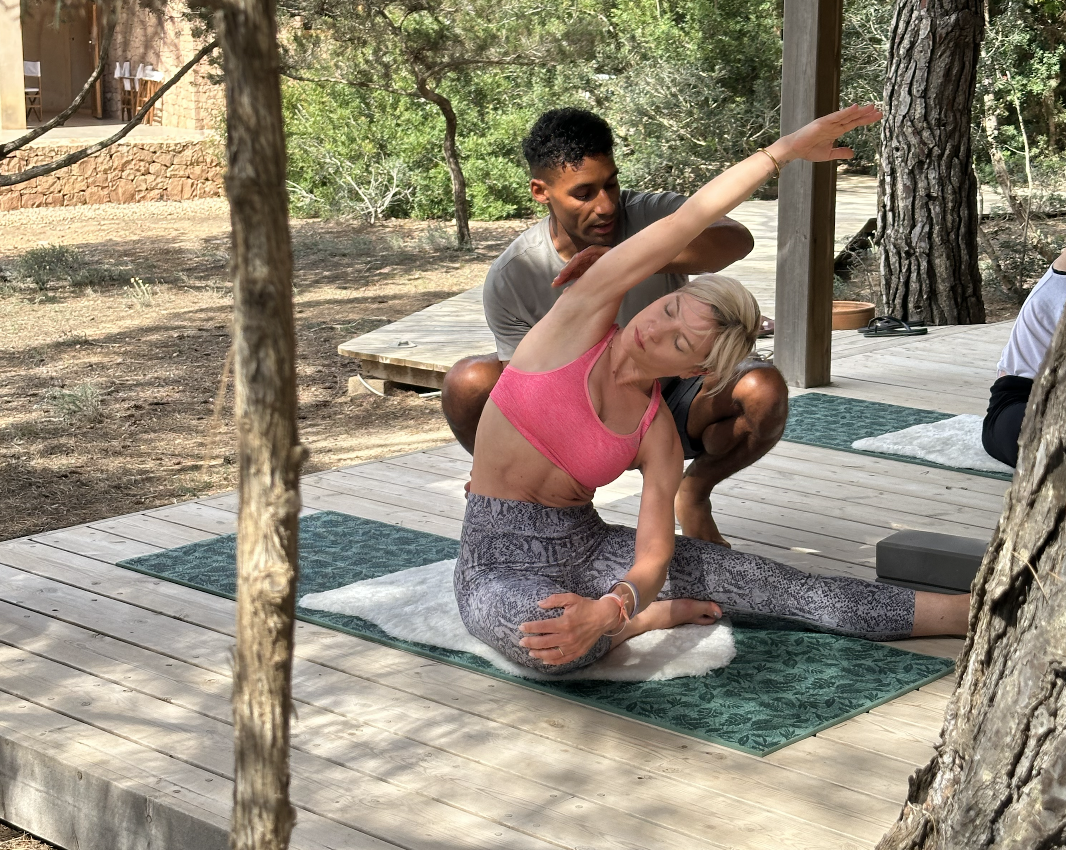 A woman practicing yoga outdoors on a wooden deck, assisted by a male instructor. She is wearing a pink sports bra and patterned leggings, sitting cross-legged with her right arm raised overhead, stretching. The environment is surrounded by trees and