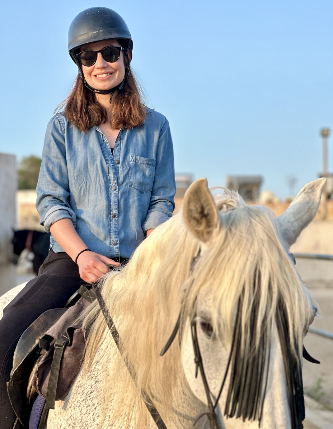 Young woman smiling, wearing sunglasses, helmet, and denim shirt, riding a white horse with a long mane.