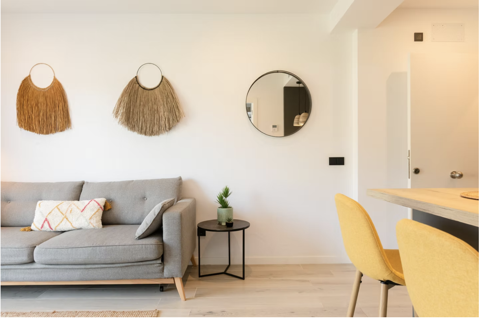 Living room with a gray sofa, a round black side table with a small potted plant, wall decorations including two woven wall hangings and a round mirror, and yellow chairs around a wooden table.