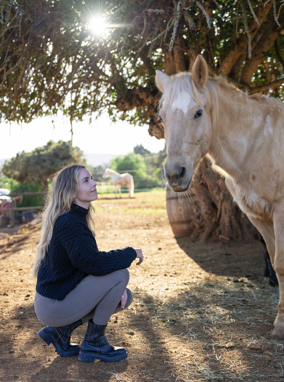 A woman squatting on the ground, facing a large light-colored horse, with another horse visible in the background under a tree and bright sunlight overhead.