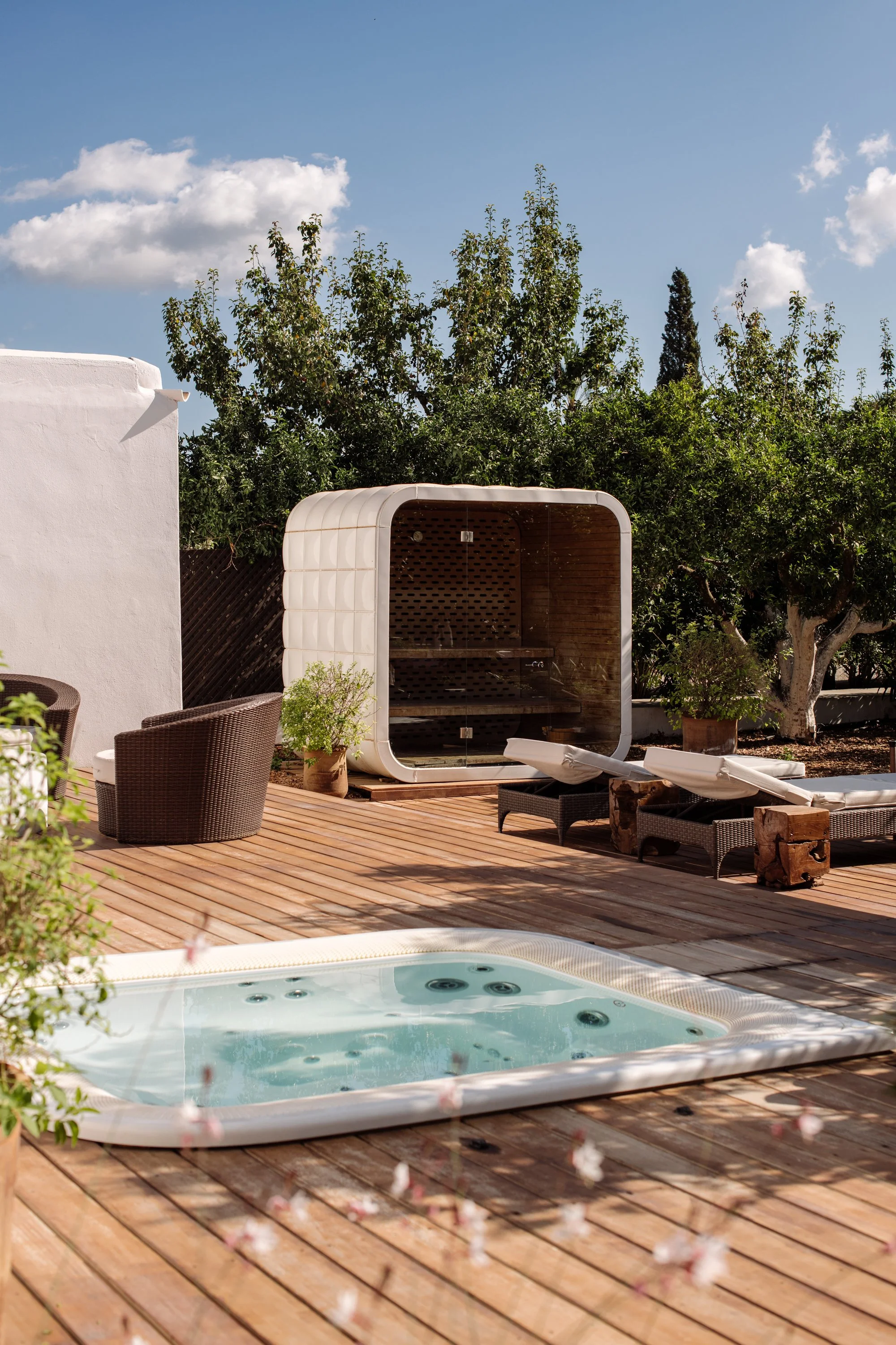 Outdoor patio with a hot tub, lounge chairs, wicker chairs, potted plants, and a small enclosed outdoor sauna on a wooden deck, with trees and clouds in the background.