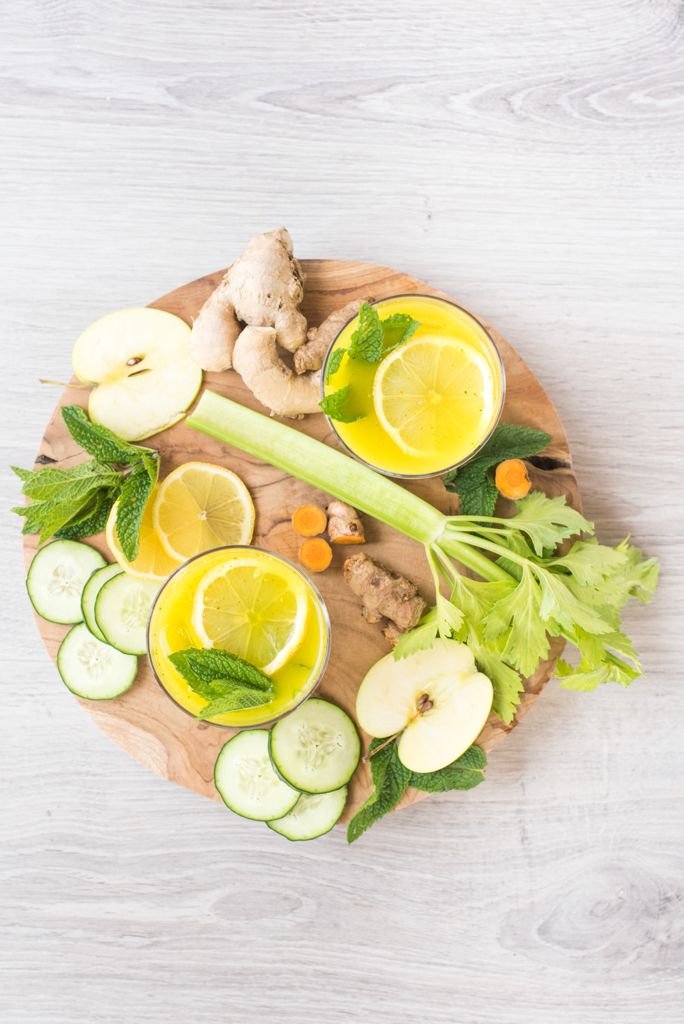 A wooden board with sliced green apples, cucumber, ginger, lemon slices, mint leaves, celery stalks, and two glasses of lemonade with lemon slices and mint.