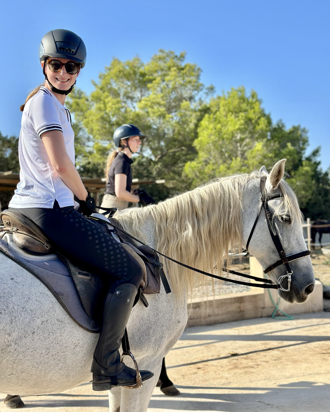Two women wearing helmets and riding attire on horseback, with trees and a clear blue sky in the background.
