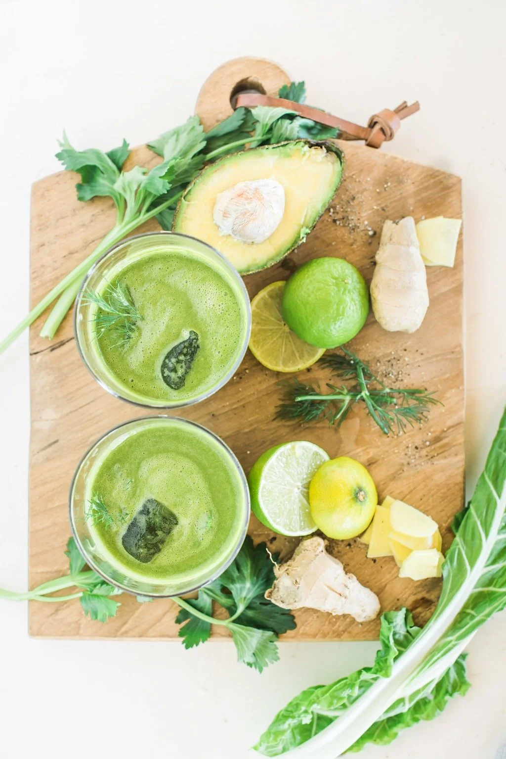 A wooden cutting board with fresh ingredients and two glasses of green juice. The ingredients include an avocado, lime, lemon, ginger, turmeric, celery, and arugula. There is a sliced lemon and a small piece of ginger on the board.