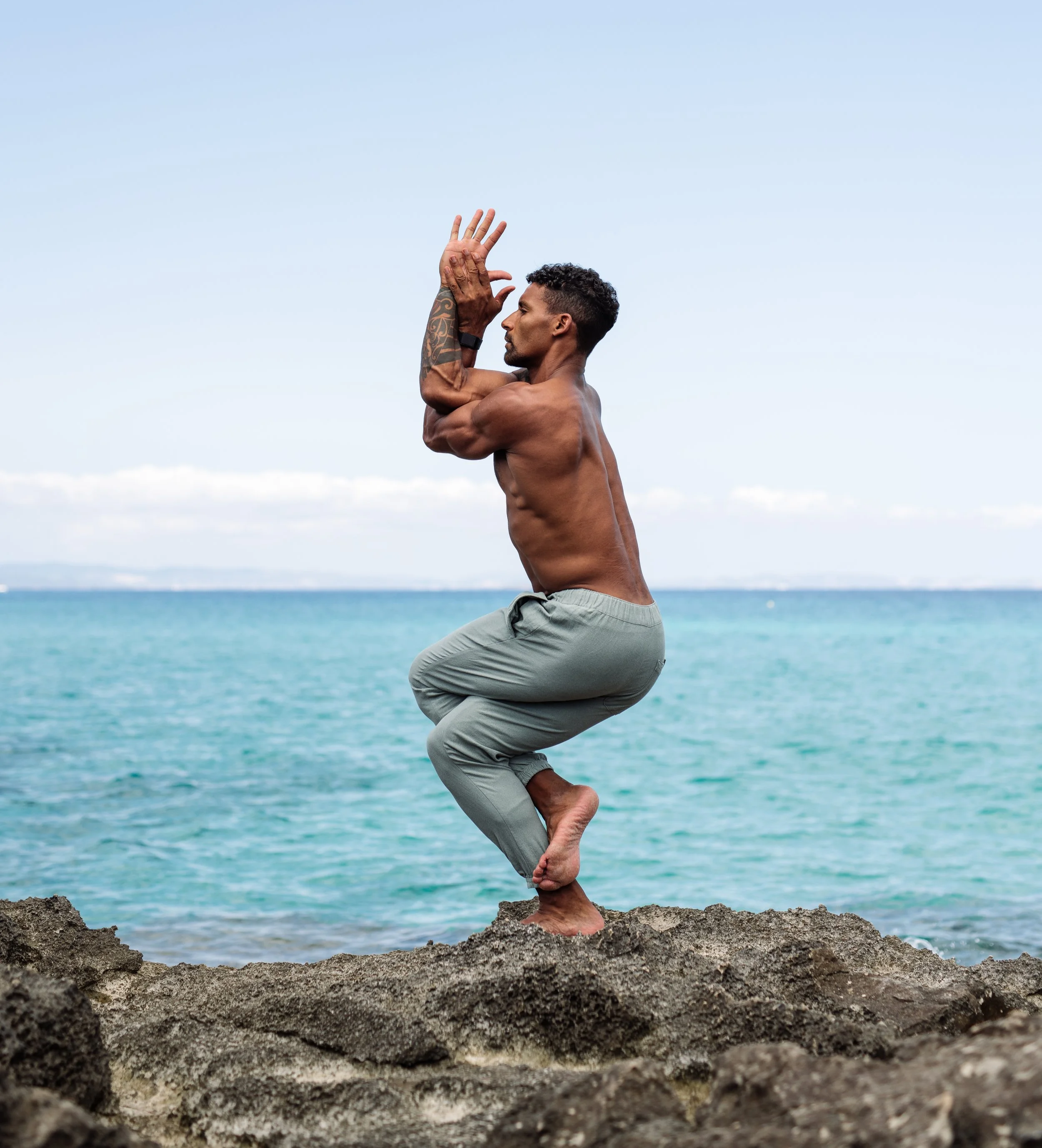 A shirtless man practicing yoga on rocks by the ocean, standing with his knees bent, arms raised with palms together near his face, with a calm sea and clear sky in the background.