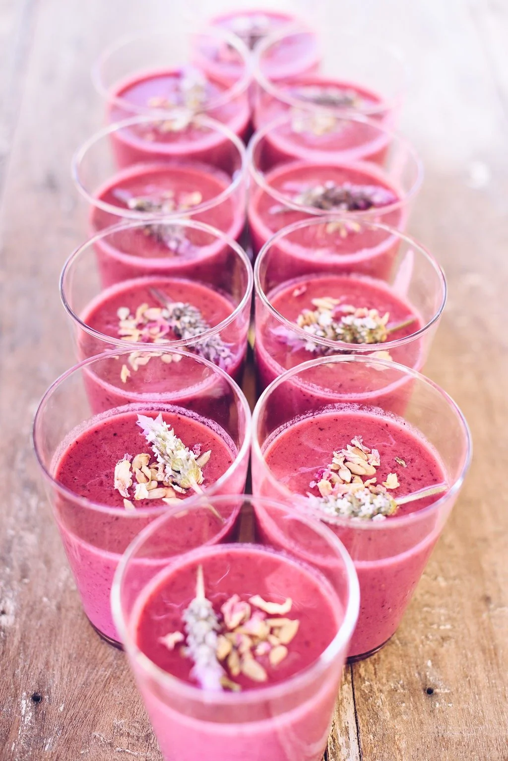 Multiple pink smoothies topped with flower and nut garnishes, arranged in rows on a wooden surface.