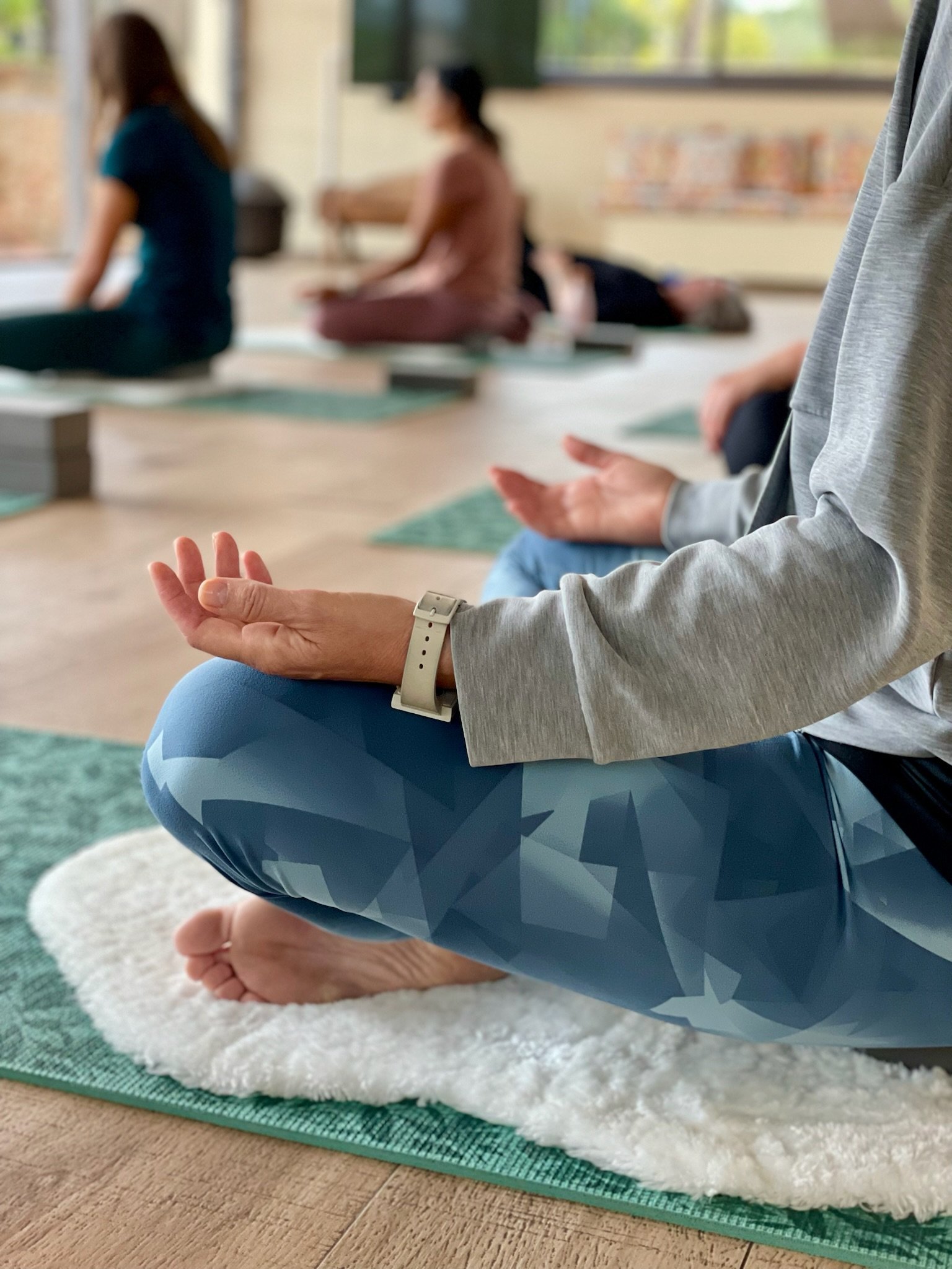 People practicing yoga indoors, seated on mats with hands resting on knees in a meditative pose.