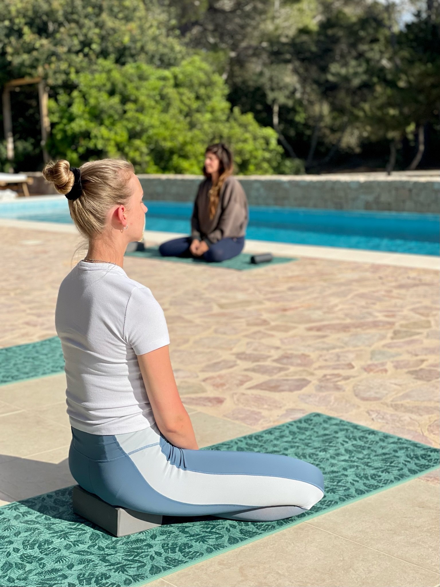 Two women practicing yoga outdoors by a pool, one sitting cross-legged on a yoga mat with hands resting on her thighs, the other sitting in a kneeling pose on a different yoga mat, surrounded by greenery.