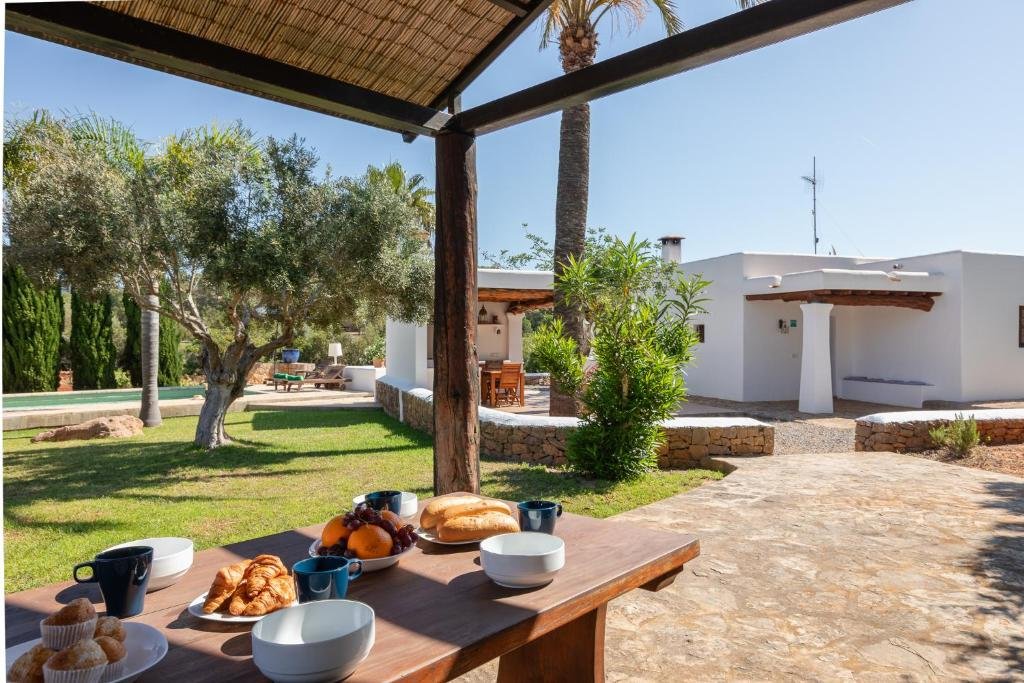 Outdoor breakfast table with croissants, bread, fruit, and cups, set under a shaded patio, with trees, greenery, and a white house with stone accents in the background.