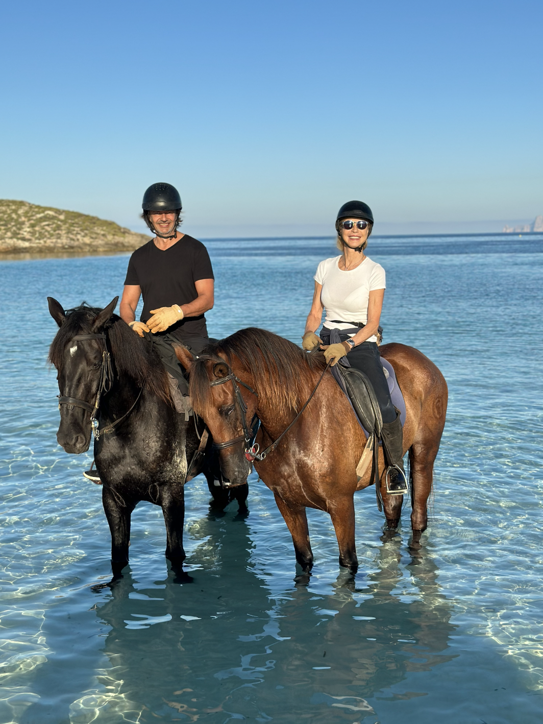 Two people riding horses in shallow clear water on a beach, wearing helmets and gloves, with a blue sky and distant land in the background.