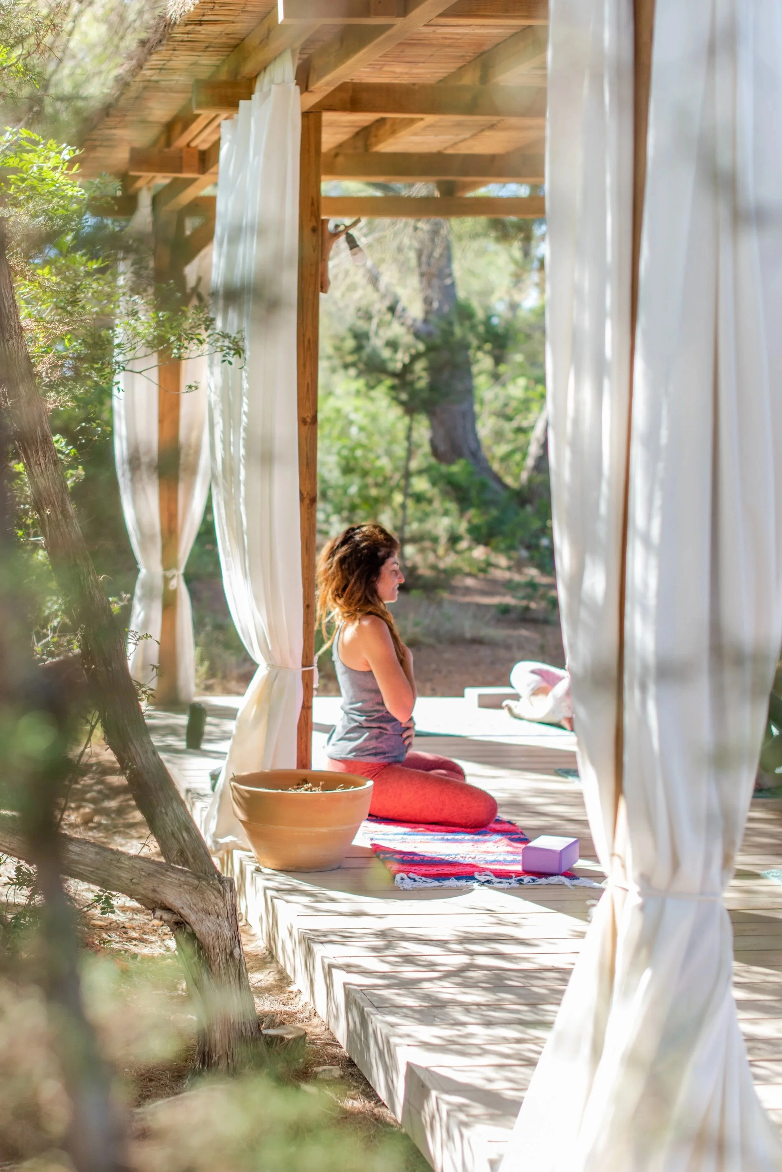 A woman practicing yoga on a wooden platform outdoors, surrounded by trees, sitting on a colorful towel, with a large pot nearby and white curtains hanging from the wooden frame.