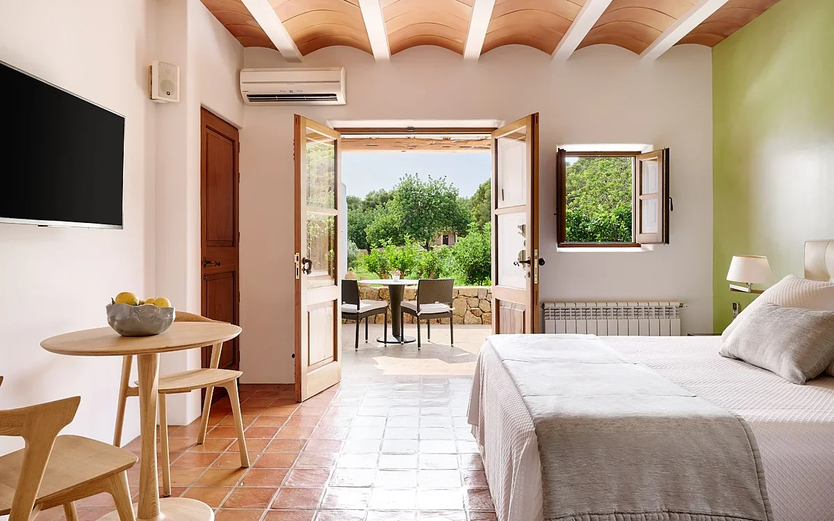 A bright hotel room with a bed, small dining table, and open patio door revealing outdoor seating and greenery.