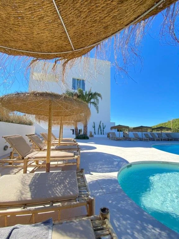 Poolside area with wooden lounge chairs under straw umbrellas, white building, palm trees, and clear blue sky.