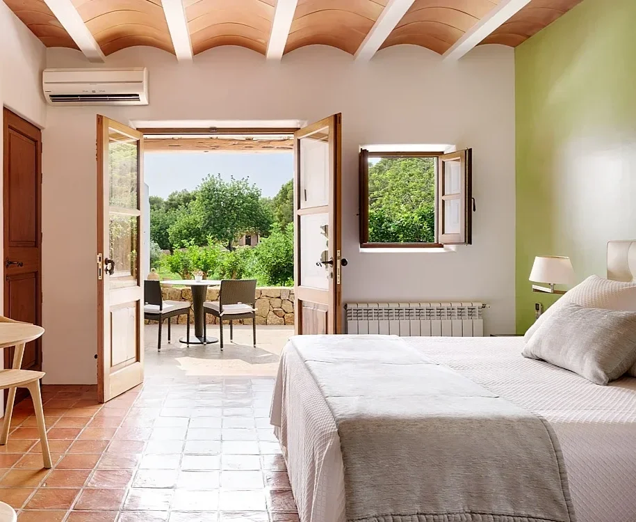Bright bedroom with a large bed, white bedding, and a small table lamp. Open double doors lead to a balcony with two chairs and a table, overlooking a lush green landscape. A window next to the doors shows trees outside, and a wall-mounted air conditioner is visible.