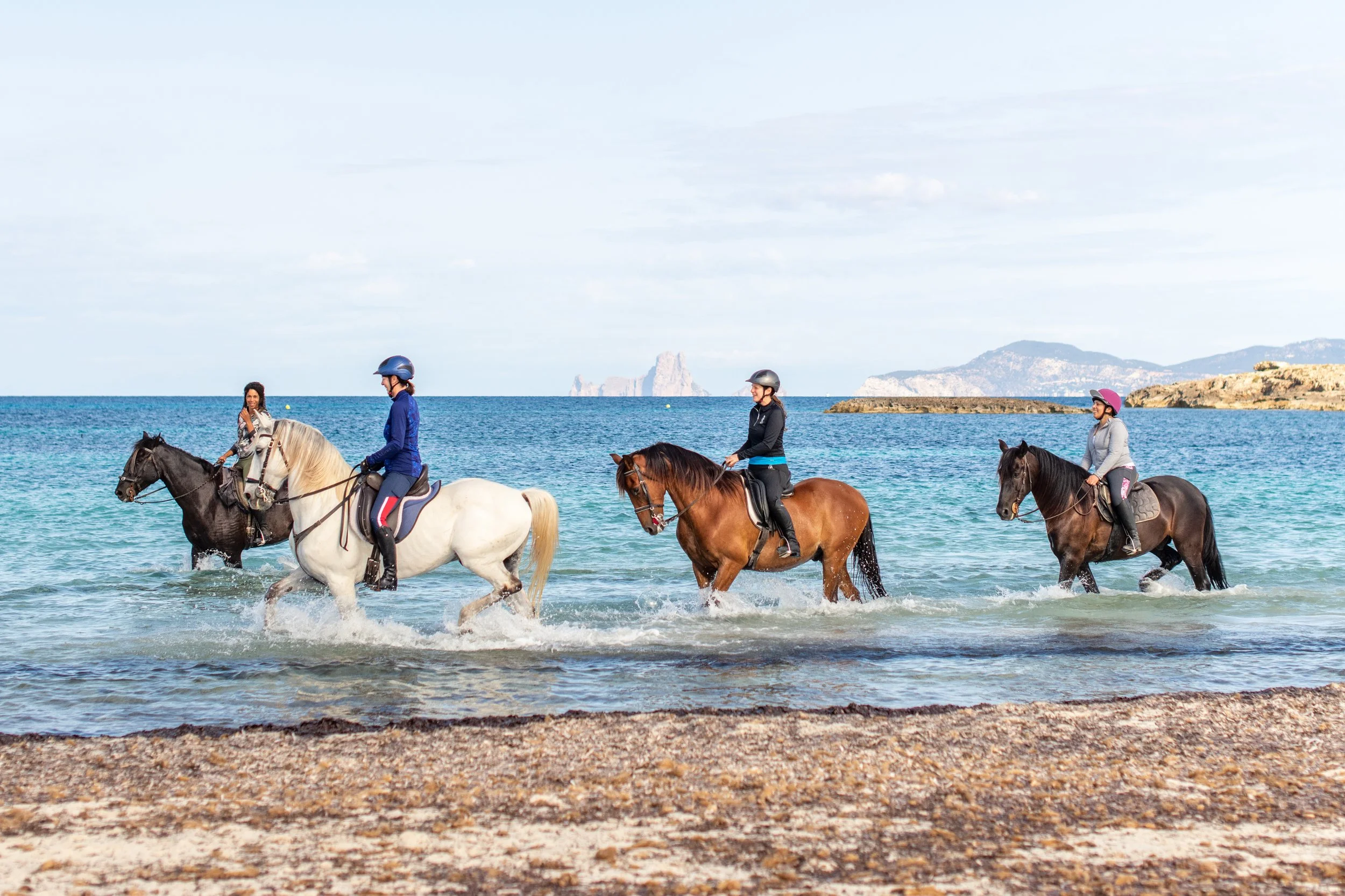 Three women riding horses in the ocean near the beach, with a rocky coastline and mountains in the background.