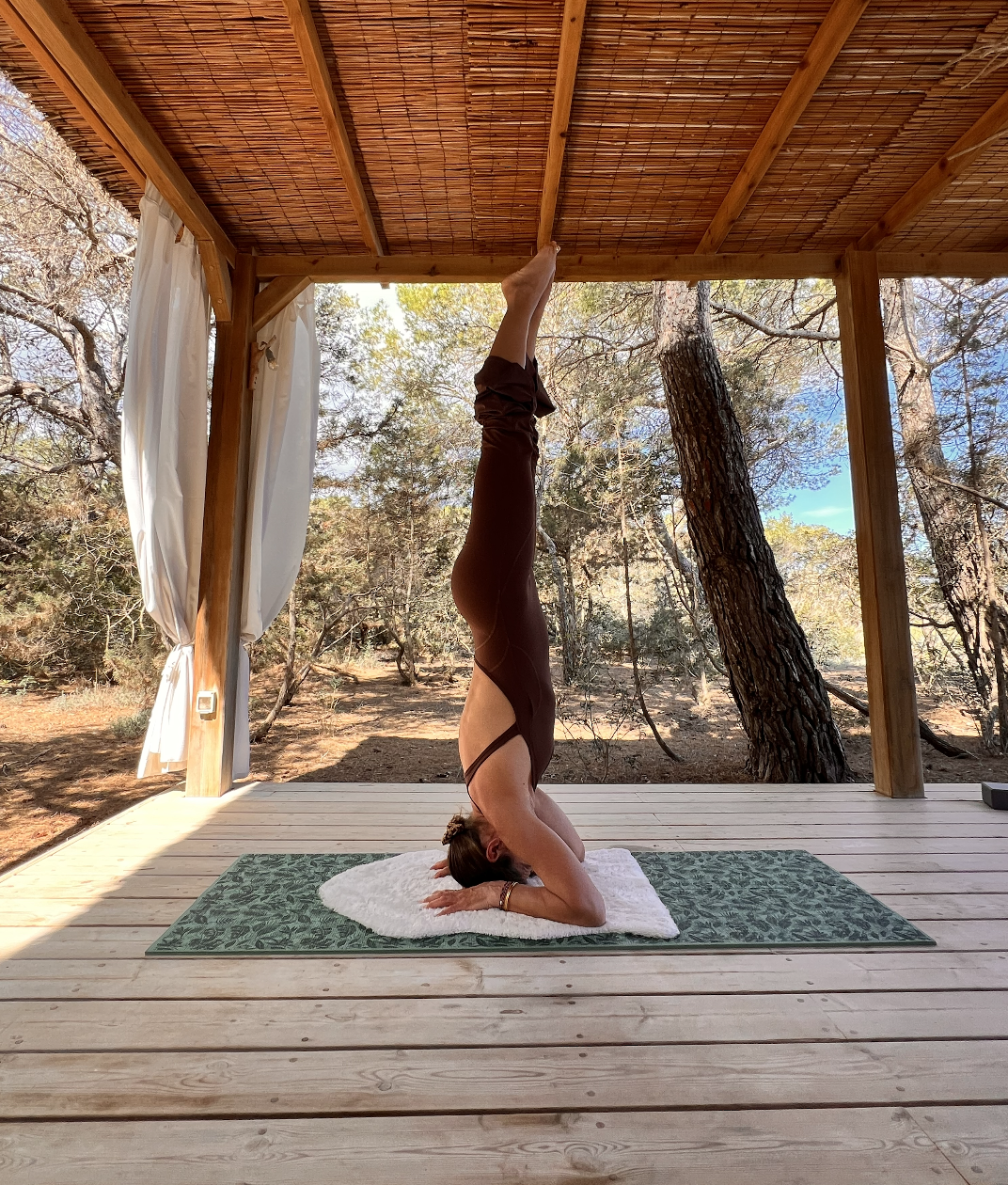 A woman practicing upside-down yoga in the headstand pose on a mat on a wooden porch surrounded by trees.