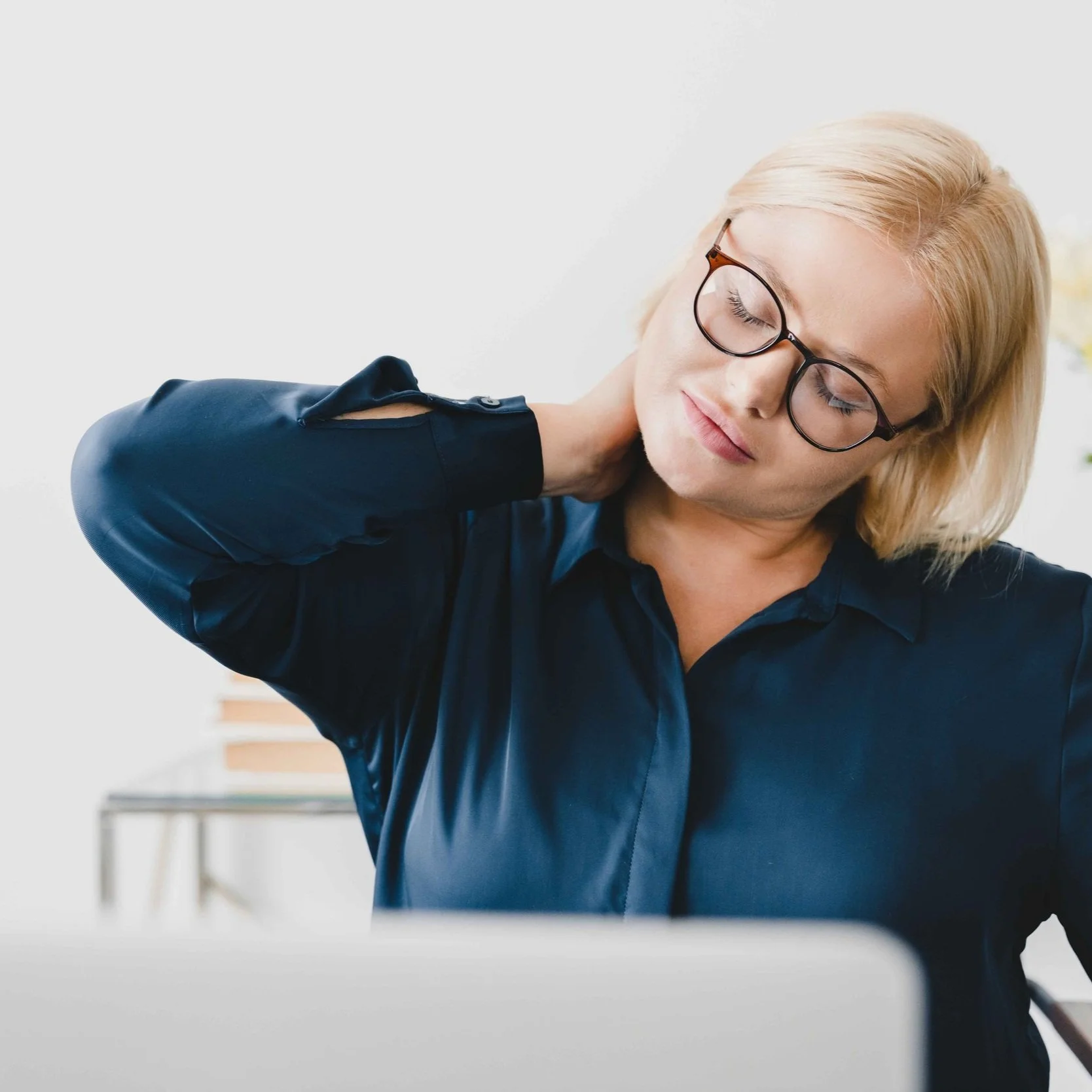 Frau mit Brille und blonder Frisur sitzt an einem Tisch, hält ihren Nacken mit der Hand und wirkt müde oder angespannt.
