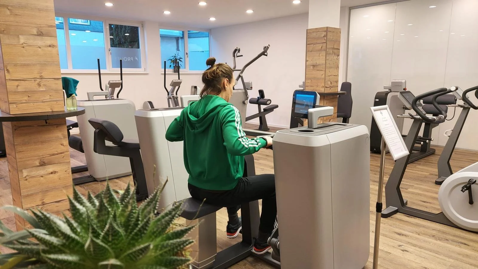 Person in grünem Trainingsanzug trainiert in einem Fitnessstudio an Kraftmaschinen, umgeben von weiteren Fitnessgeräten und einer Pflanze im Vordergrund.