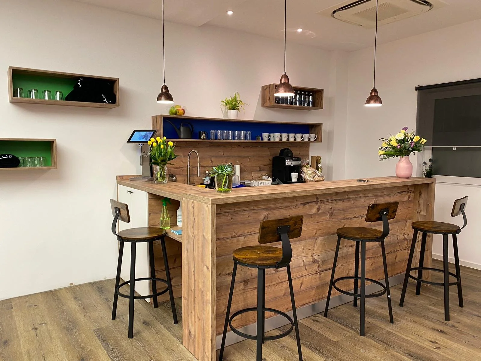 Moderne Kaffeebar mit Holztresen, vier Barhockern und dekorativen Blumenvasen. An den Wänden befinden sich Regale mit Gläsern und Tassen. Die Bar ist mit Hängelampen beleuchtet.