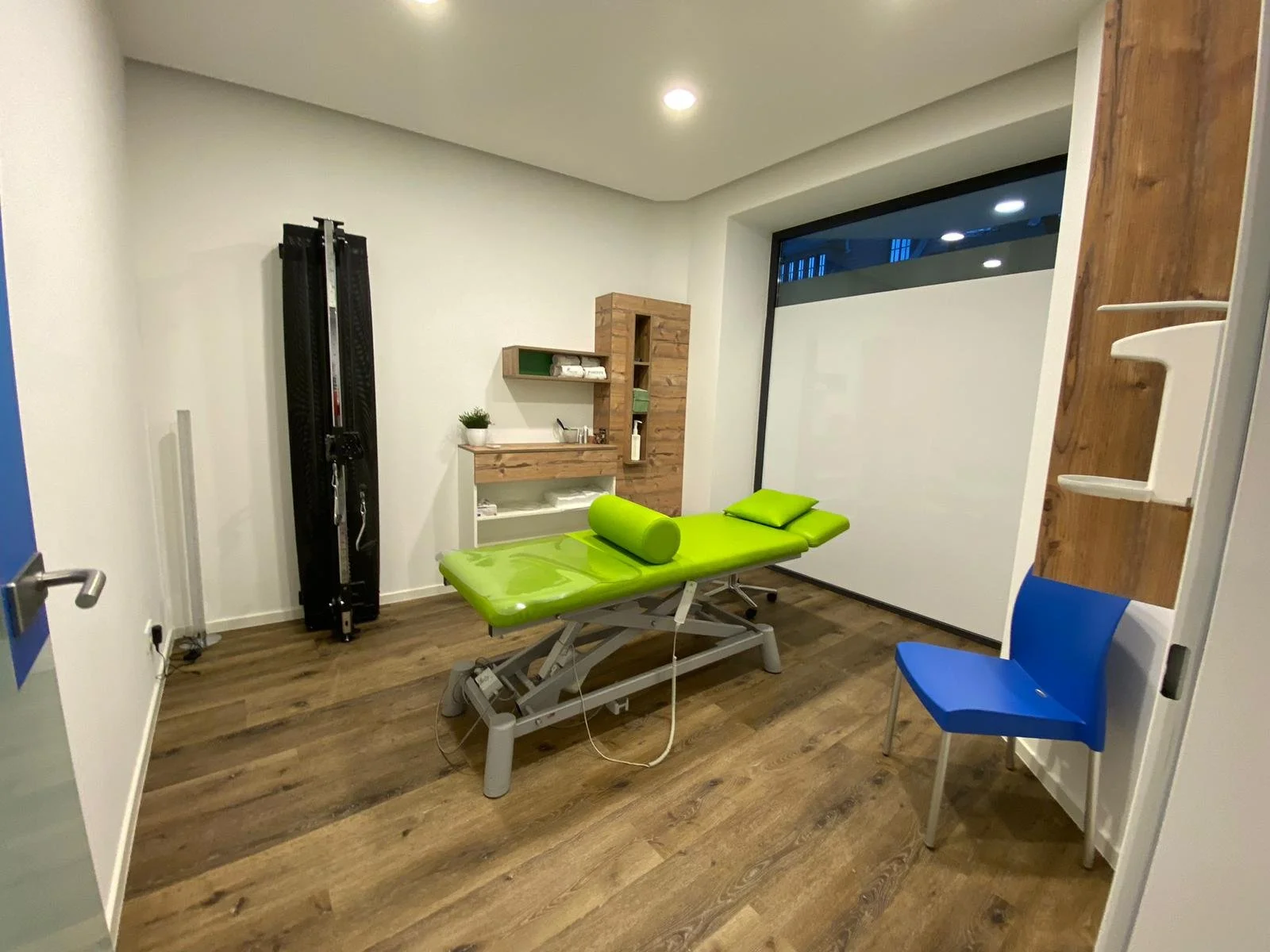 Physiotherapieraum mit grünem Behandlungstisch, blauer Stuhl, Holzboden und Schrank.