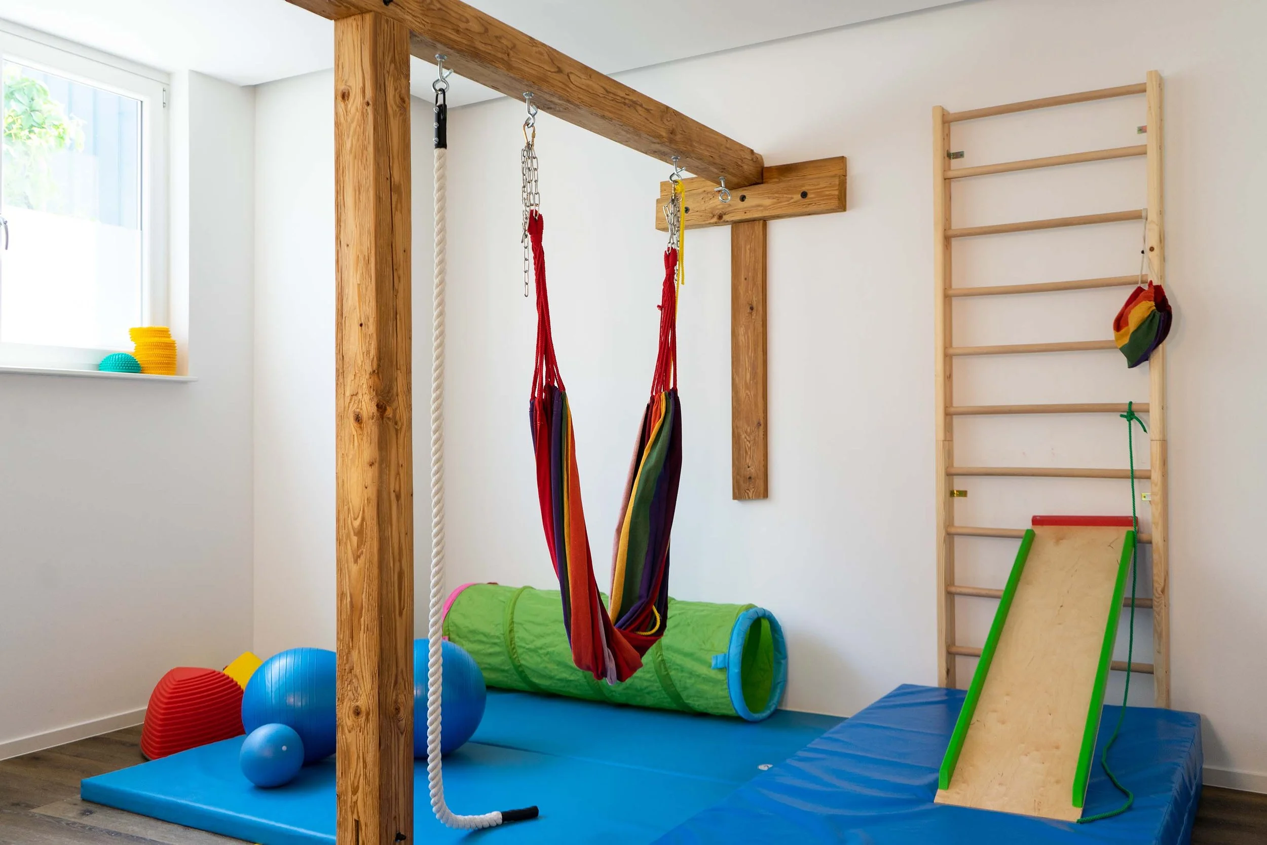 Innenraum eines Kinder-Fitnessraums mit Hängematte, Kletterseil, Sprossenwand, Tunnelspielzeug und verschiedenen bunten Fitnessbällen.