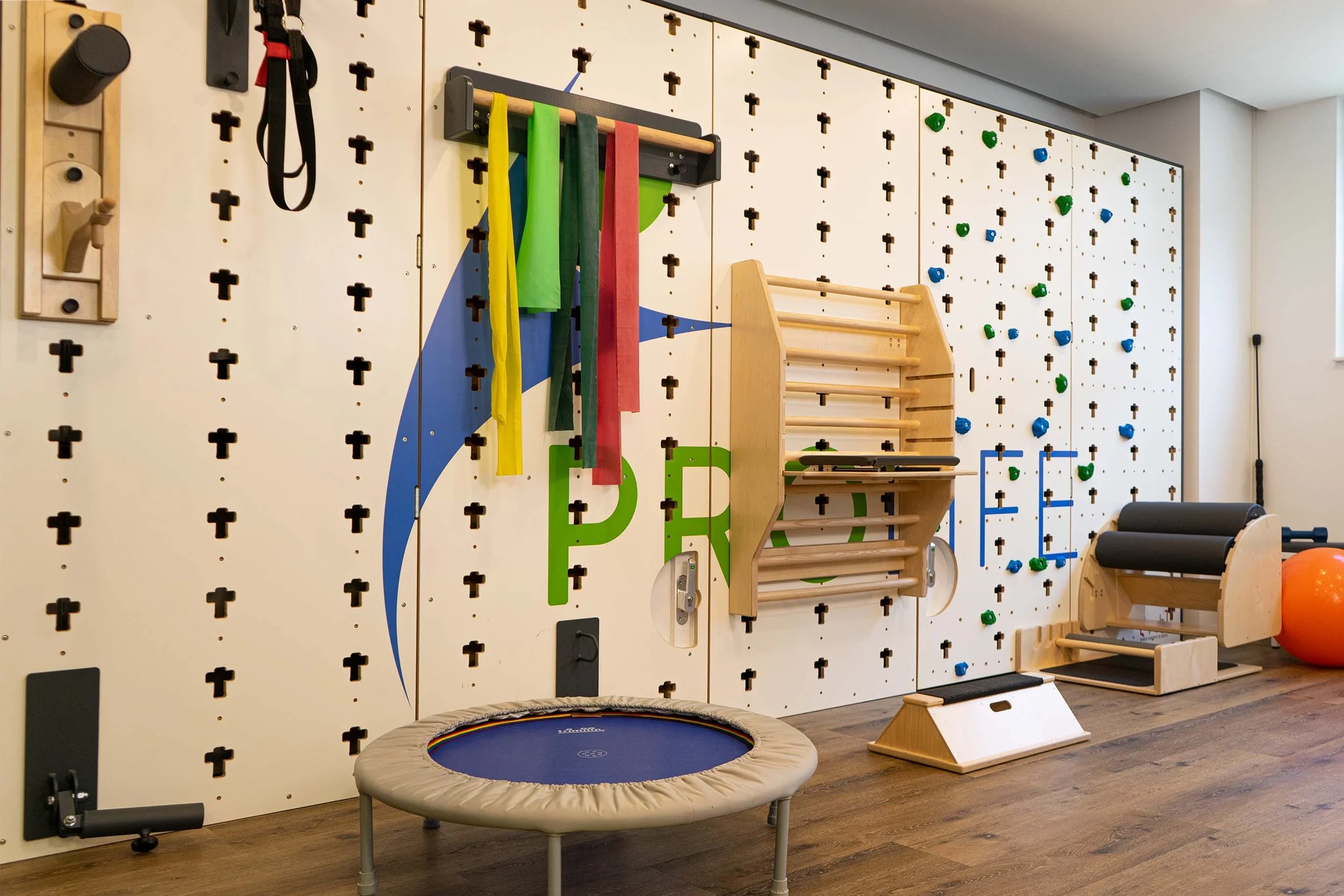 Fitnessraum mit weißen Wänden, Kletterwand, bunten Widerstandsbändern, Trampolin und verschiedenen Fitnessgeräten auf Holzboden.