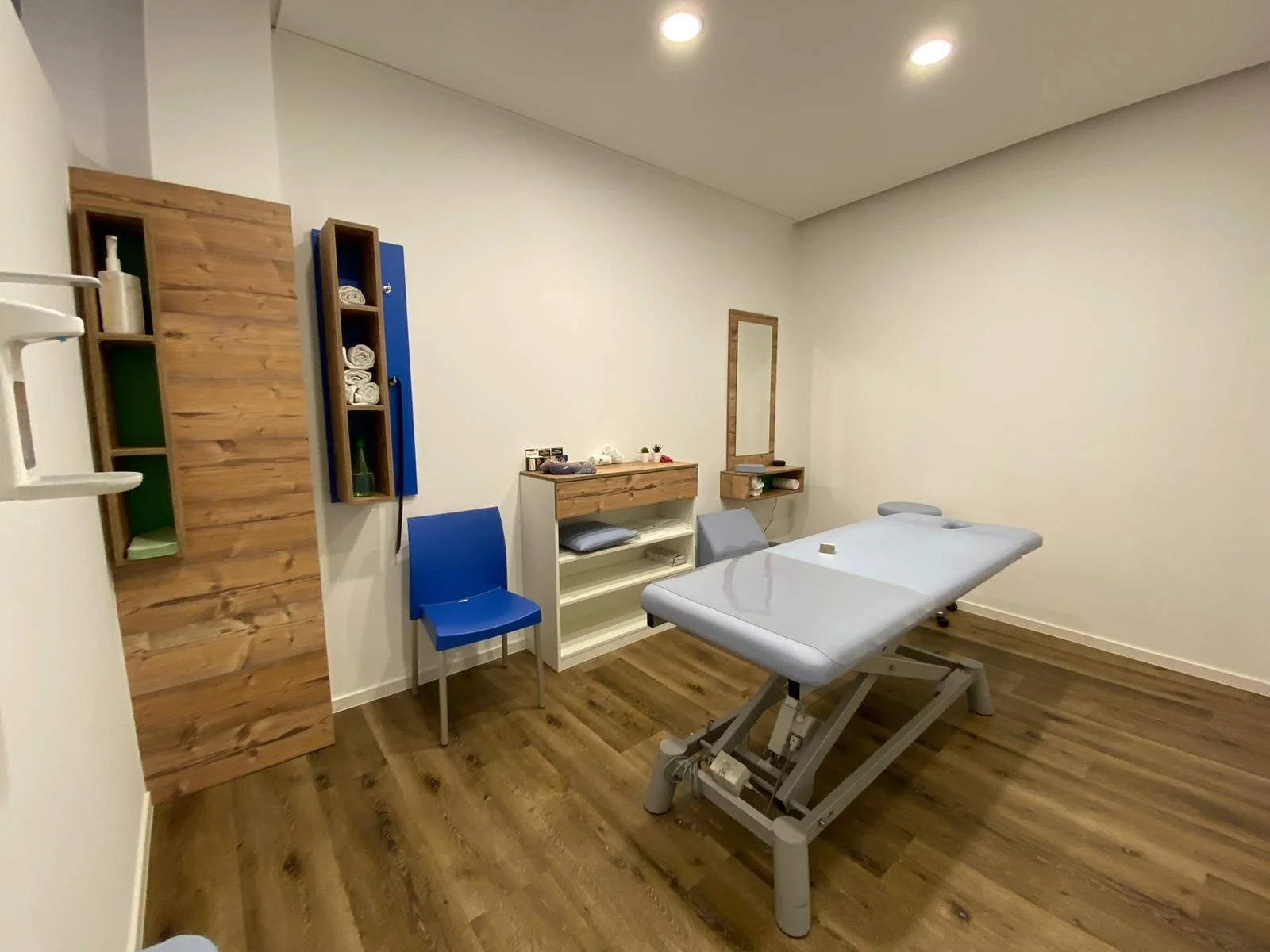 Raum mit einem Massagetisch, blauen Stuhl, Regalen und Holzfußboden.