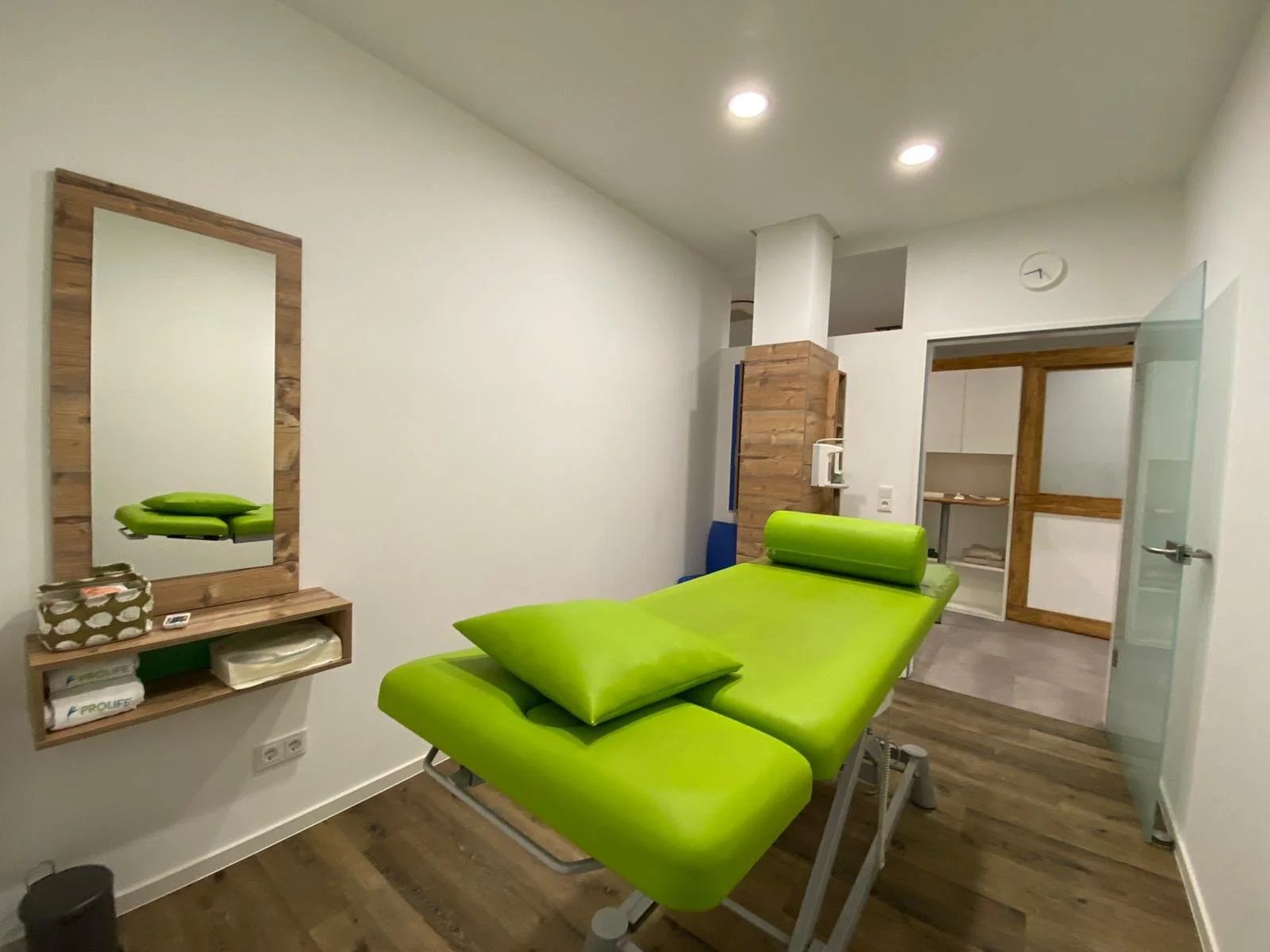Ein Behandlungsraum mit einer grünen Behandlungsliege, einem Wandspiegel aus Holz, Regalen mit Utensilien und einer offenen Tür zu einem anderen Raum.
