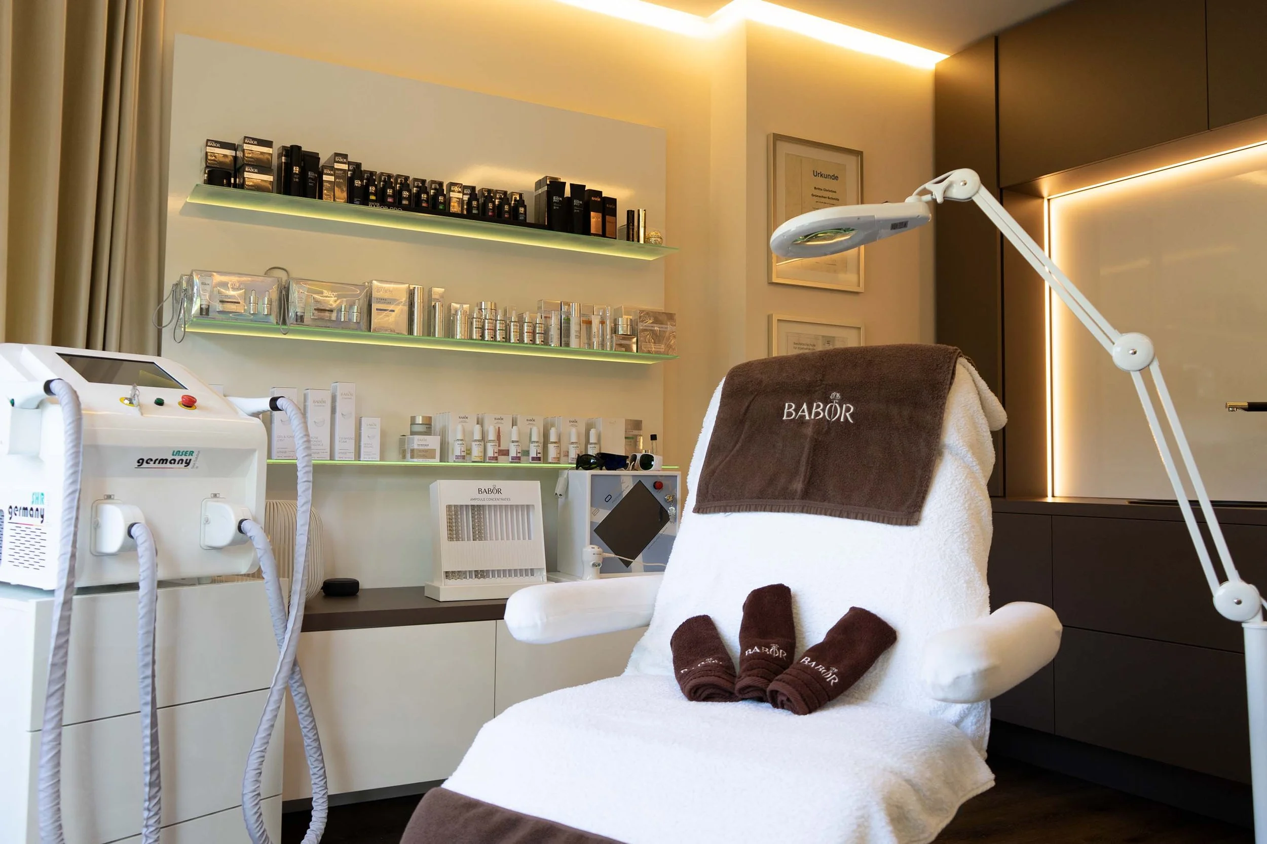 Innenraum eines Kosmetikstudios mit einem Behandlungsstuhl, umgeben von Pflegeprodukten und Geräten. Ein Regal zeigt verschiedene Pflegeartikel. Ein Gerät mit Schläuchen steht links vom Stuhl.