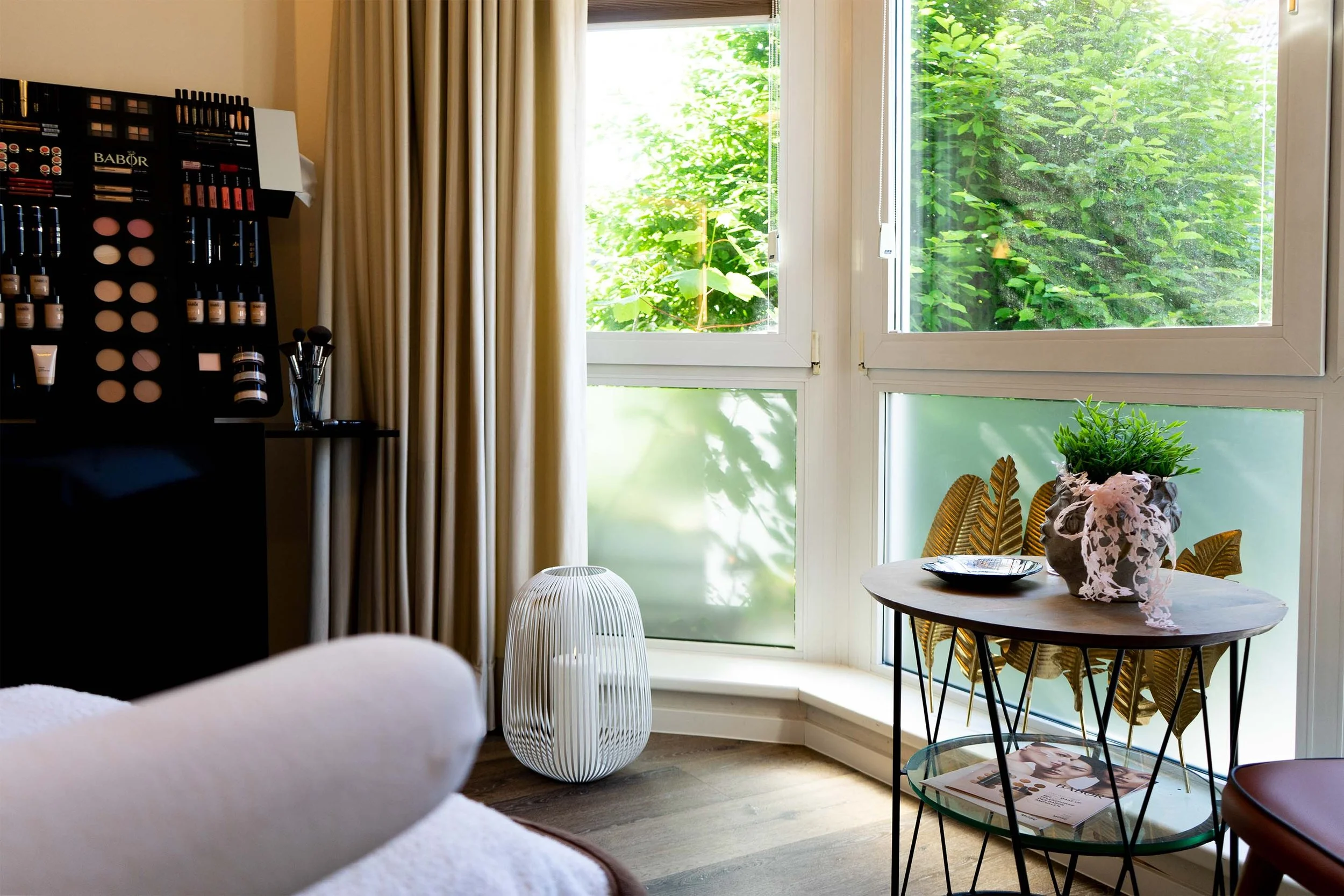 Innenraum eines Kosmetikstudios mit Make-up-Produkten, Tisch mit Pflanze, Zeitschriften, Fenster mit Ausblick auf Bäume.