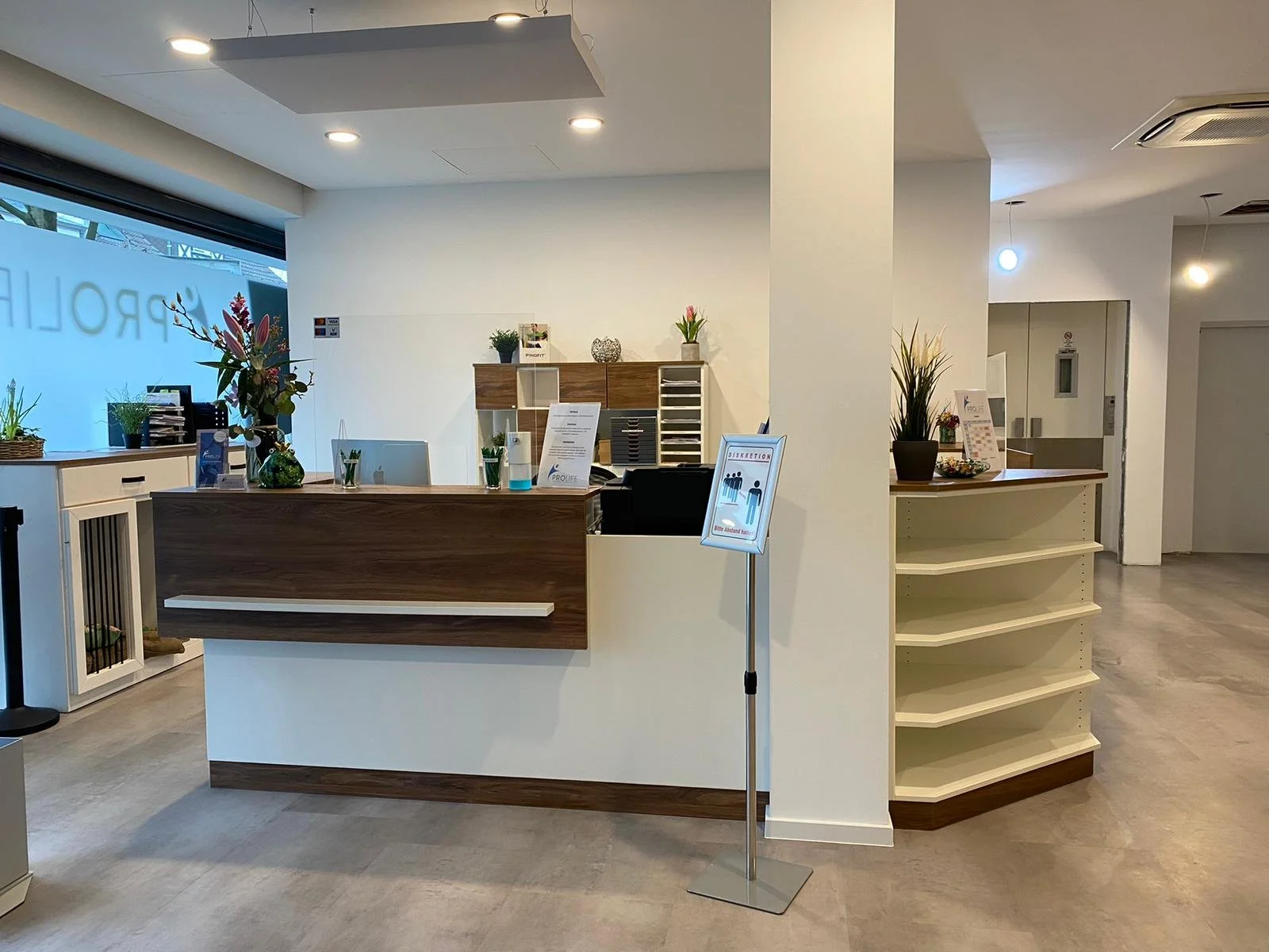 Empfangsbereich mit Tresen aus Holz und weißer Wand dahinter. Auf dem Tresen stehen Blumen und Dekorationen. Im Hintergrund sind Regale und weitere Pflanzen. Ein Schildständer ist neben dem Tresen platziert. Der Boden ist aus grauem Material.