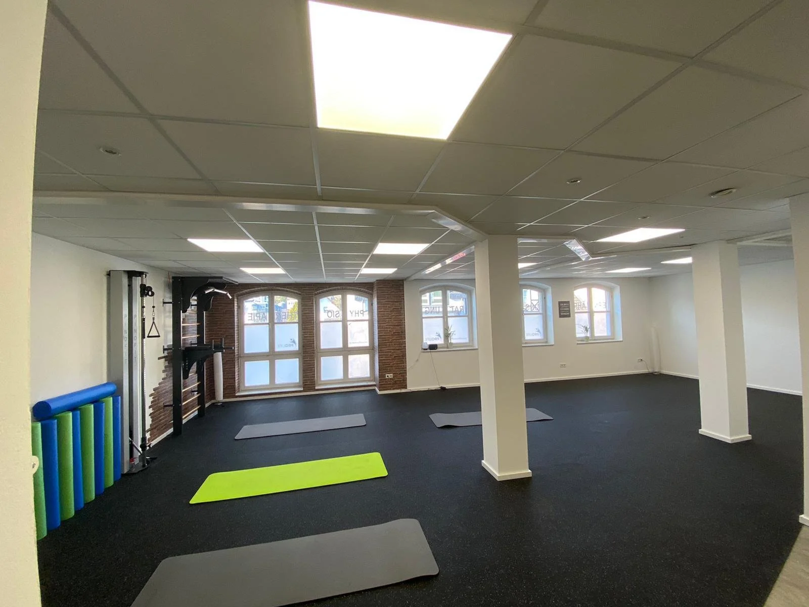 Fitnessstudio-Raum mit Gymnastikmatten, Trainingsgeräten und Fenstern, die natürliches Licht hereinlassen.