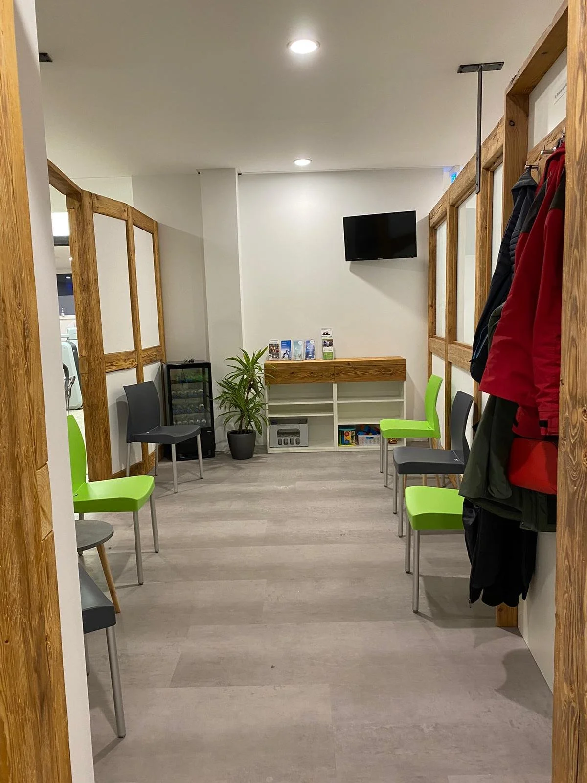 Wartezimmer mit mehreren Stühlen, teils grün und teils schwarz, entlang der Wände. An der Wand hängt ein Fernseher. Ein paar Broschüren sind auf einem Regal neben einem kleinen Kühlschrank platziert. Eine Pflanze steht daneben. Es hängen Jacken an der Wand.