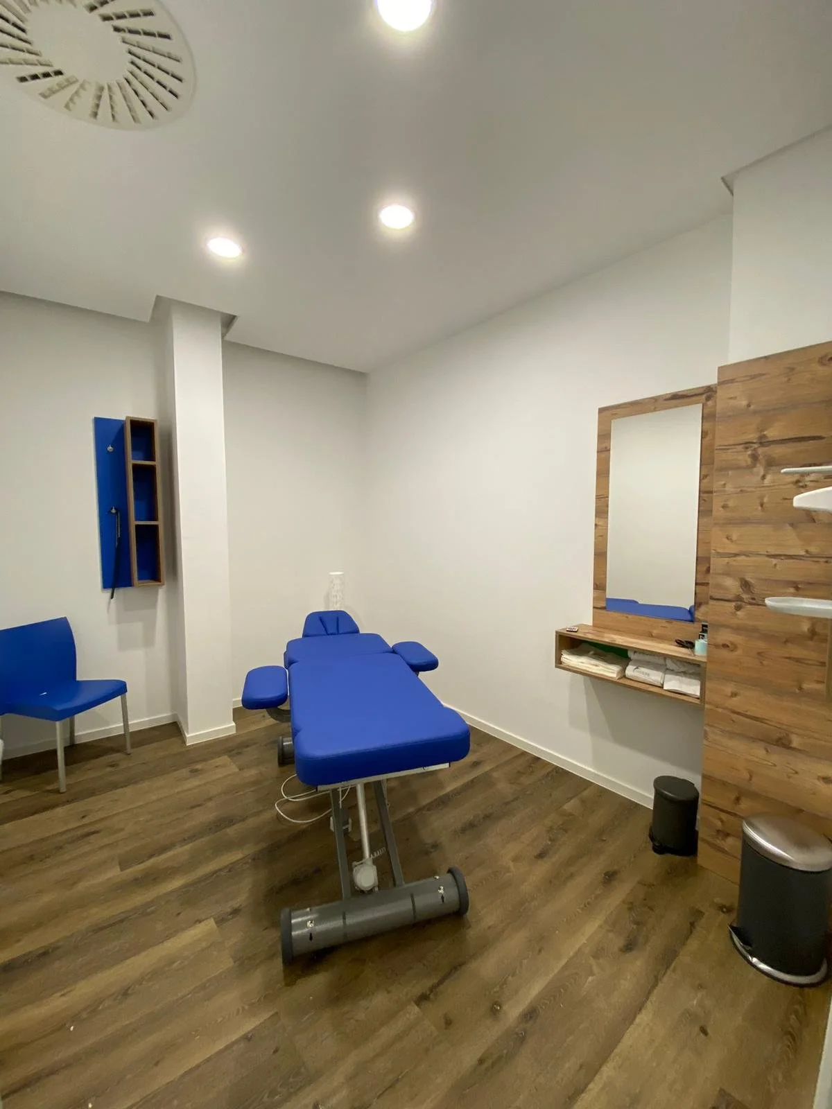 Behandlungsraum mit blauer Behandlungsliege, Holzfußboden, Wandspiegel und Regalen. Beleuchtung mit Deckenspots.