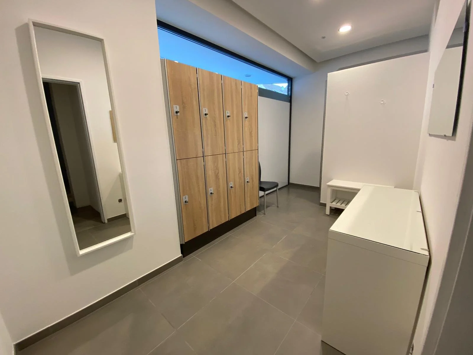 Umkleideraum mit Holzspinden, einem Spiegel an der Wand, einer Bank und Fliesenboden.