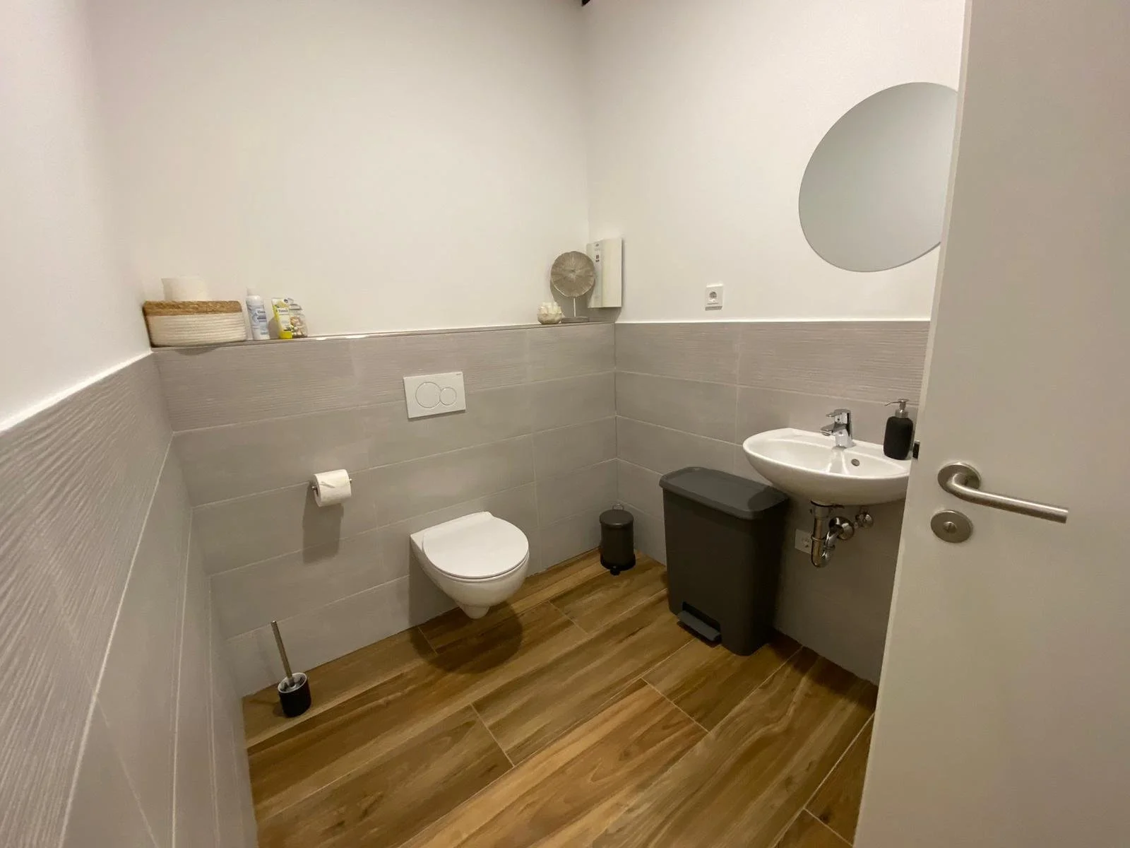 Modernes Badezimmer mit Wandtoilette, Waschbecken und Mülleimer. Graue Wandfliesen und Holzboden. Ein runder Spiegel hängt über dem Waschbecken.