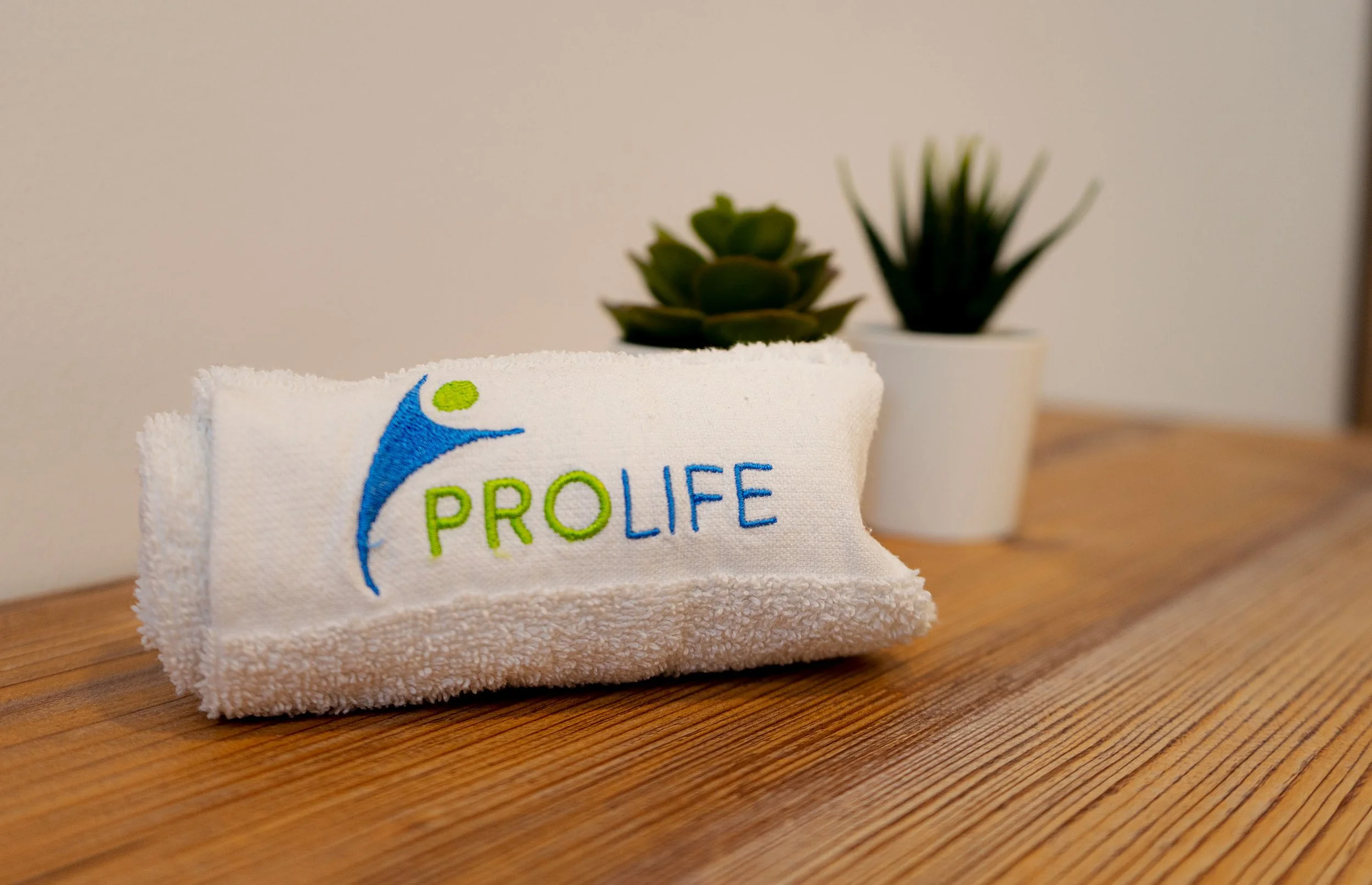 Ein weißes Handtuch mit dem Logo und Schriftzug "PROLIFE" darauf, auf einem Holztisch, mit zwei kleinen Pflanzen im Hintergrund.